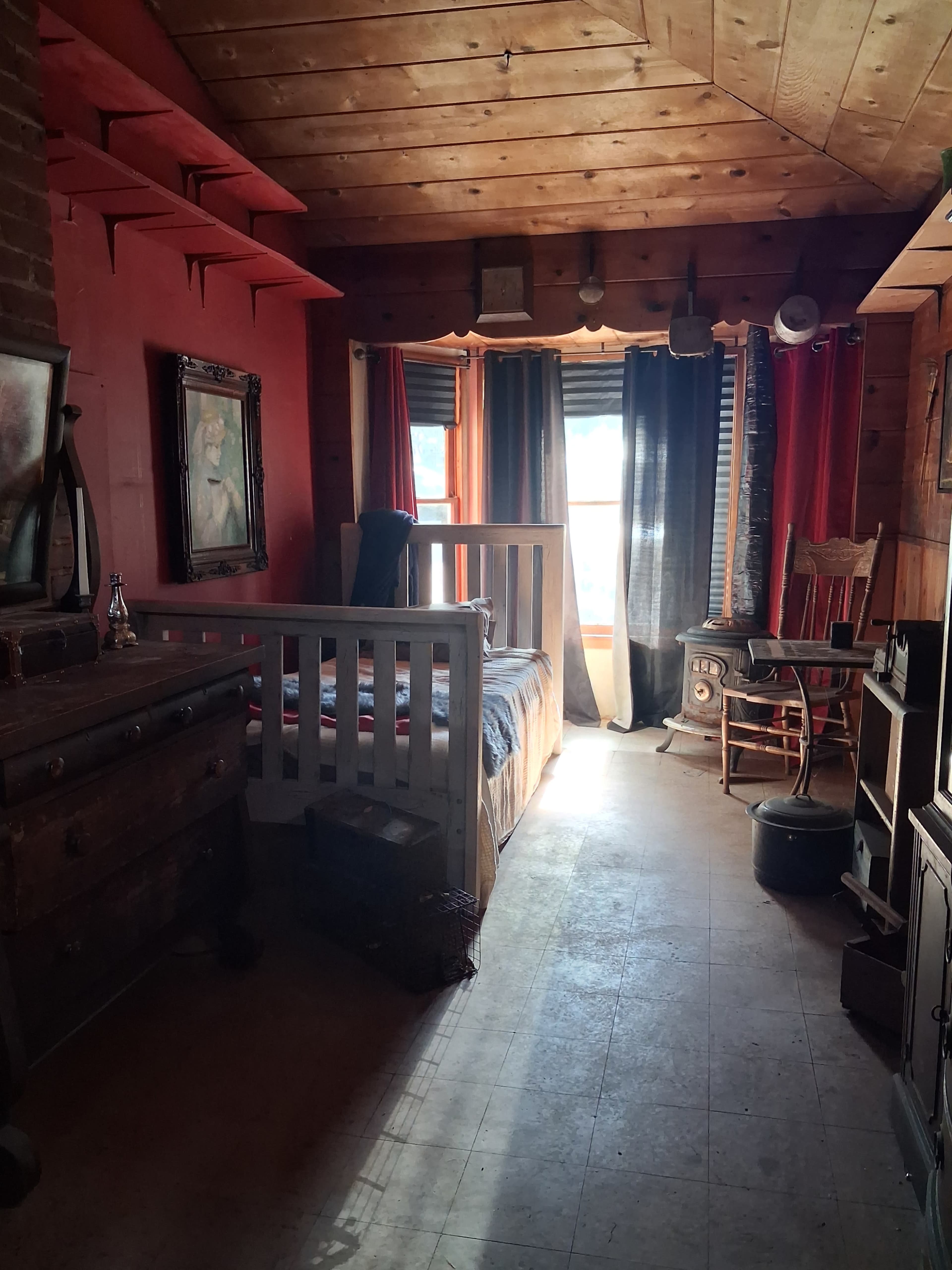 The image shows a rustic bedroom with wooden walls, a bed, a rocking chair, and a window draped with curtains.
