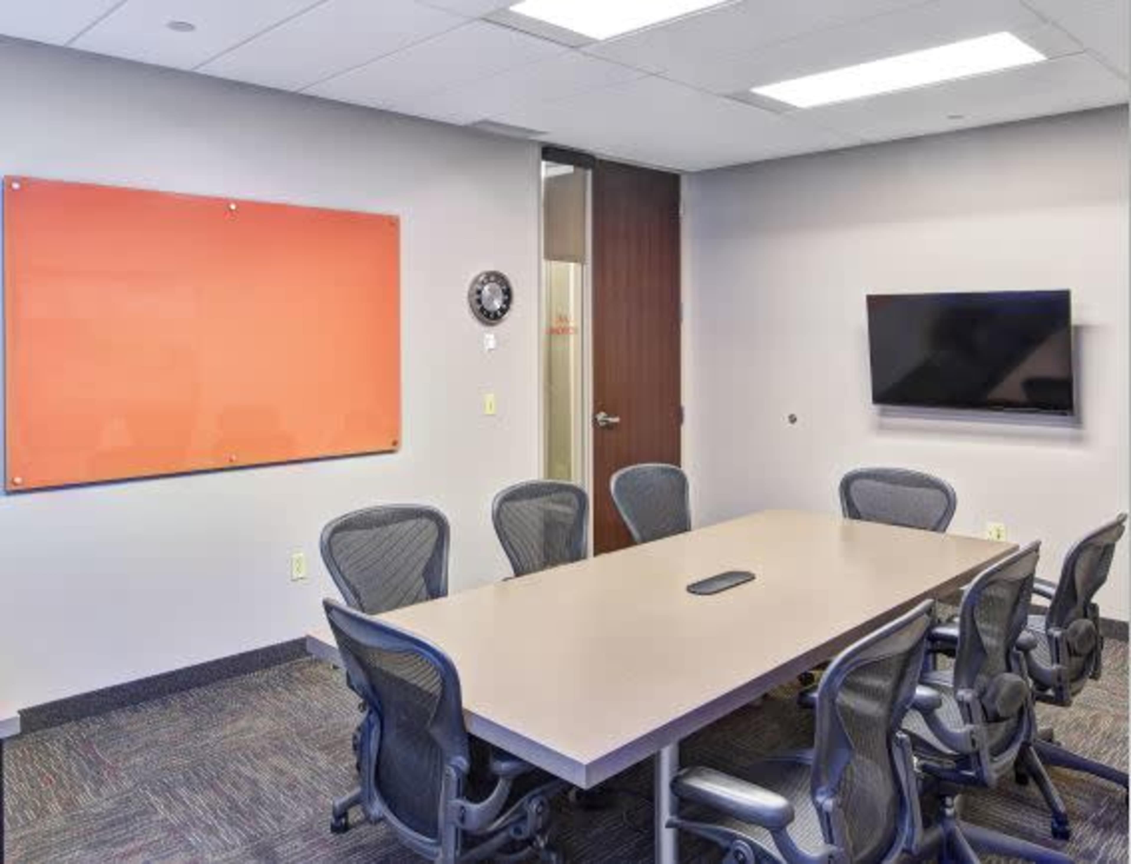 The image shows a conference room with a long table, ergonomic chairs, a whiteboard on the wall, and a wall-mounted television.