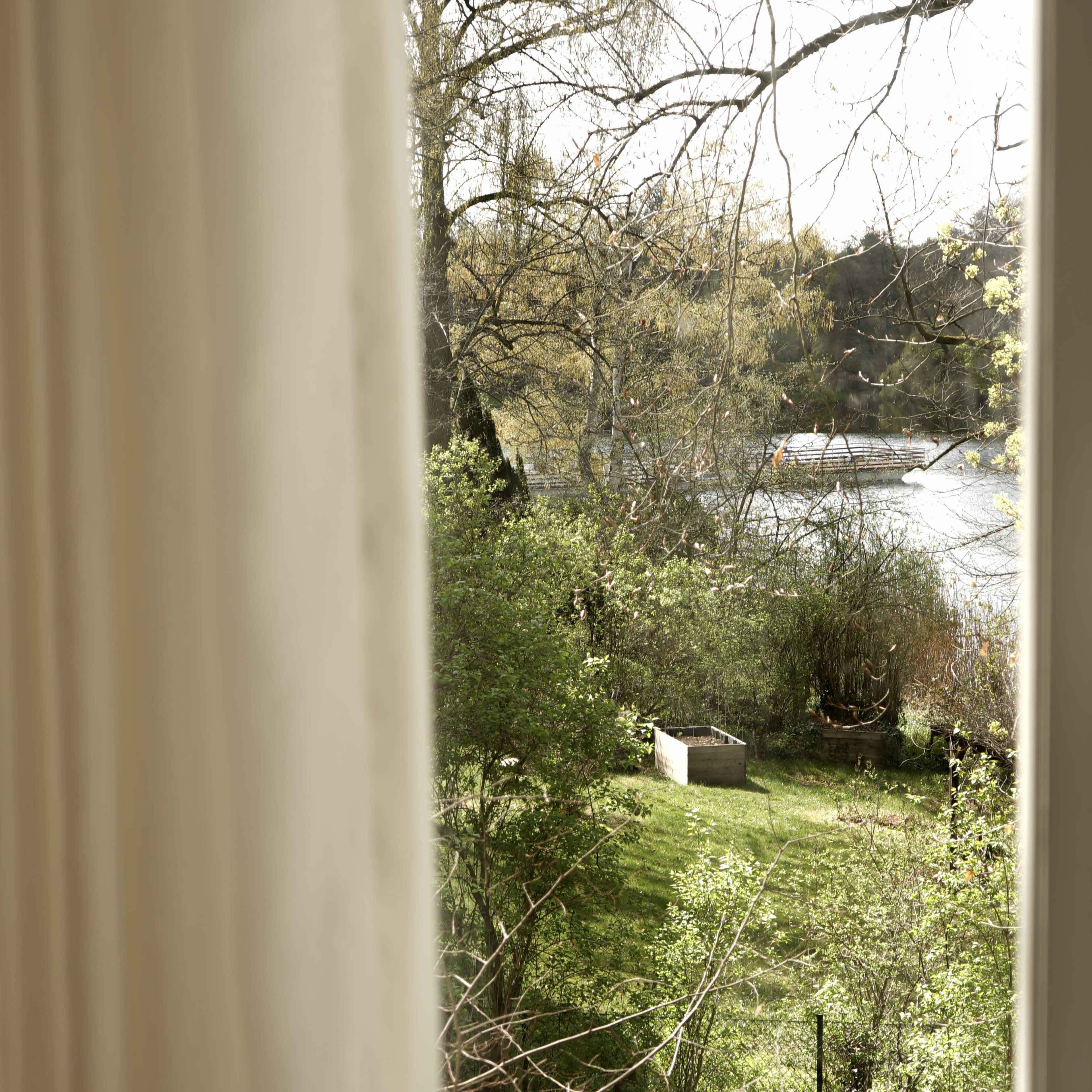 The image shows a view from a window overlooking a green garden area with a small body of water and trees in the background.
