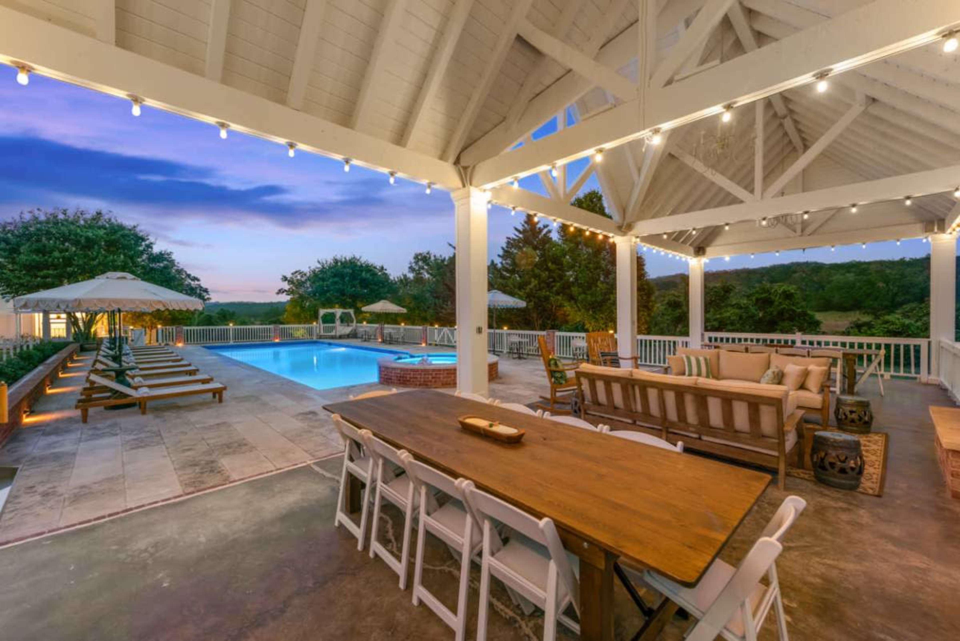 The image shows an outdoor patio area with a dining table, lounge seating, a swimming pool, and lounge chairs surrounded by greenery at dusk.