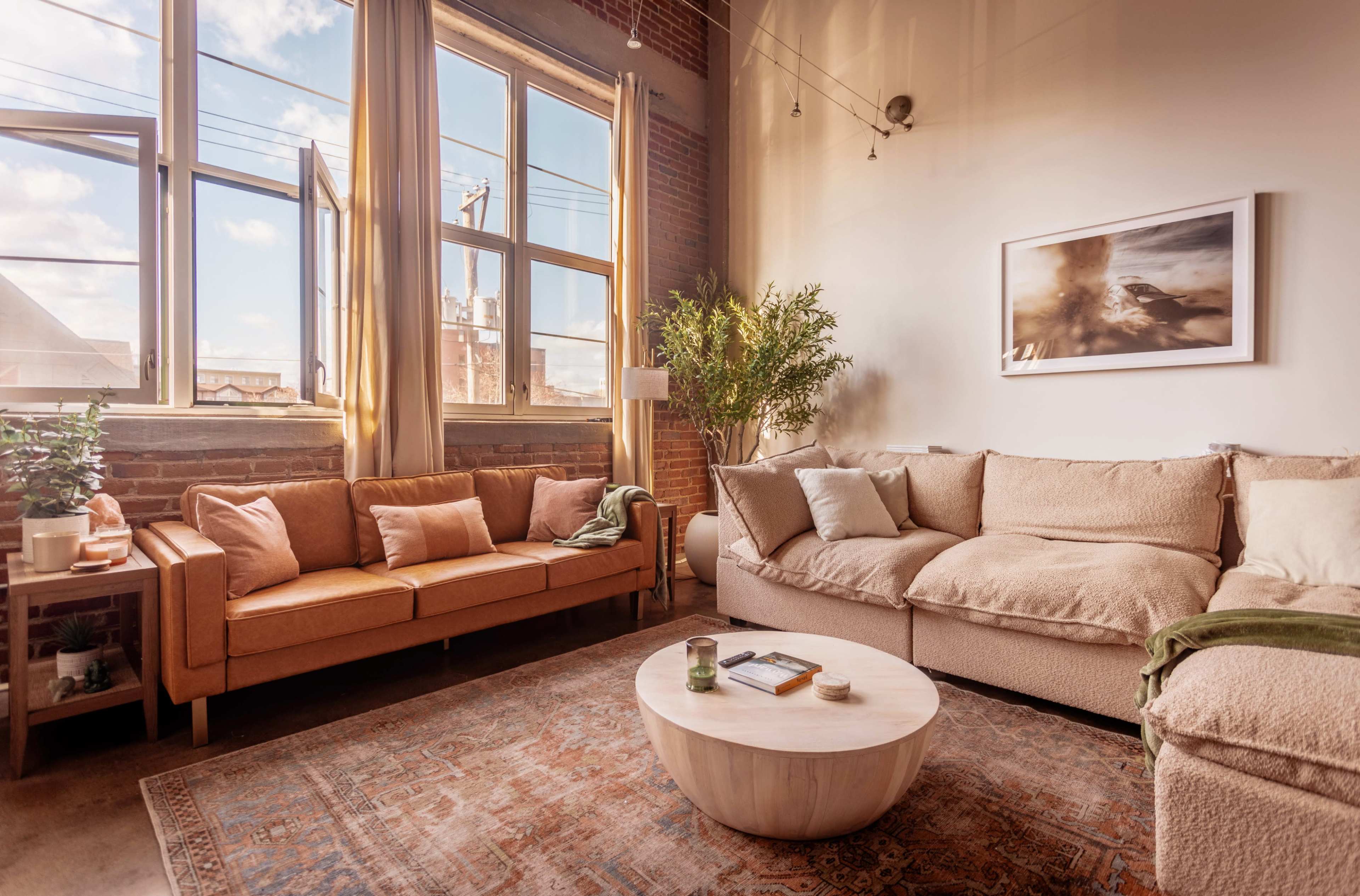 The room features a cozy seating area with a beige sectional sofa, an orange couch, a round wooden coffee table, and large windows allowing natural light to flood the space.