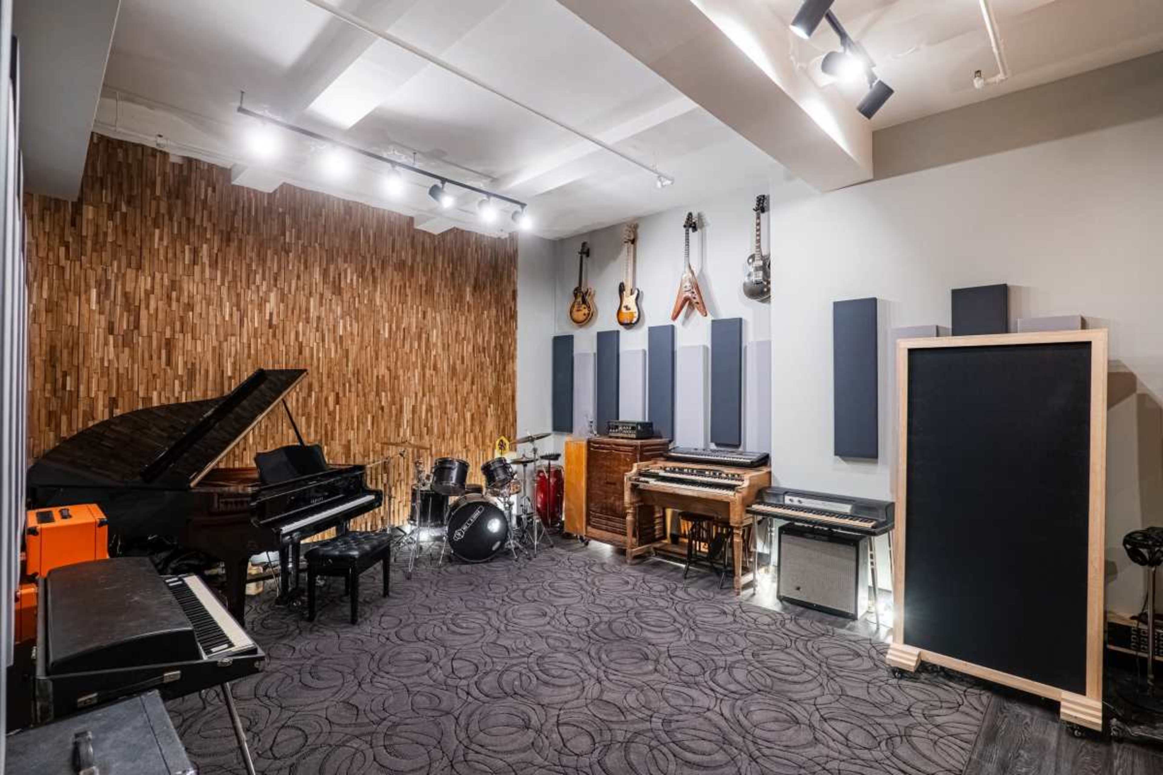 The image shows a music studio filled with various instruments, including a grand piano, guitars, a drum set, and several amplifiers arranged against a wooden wall.