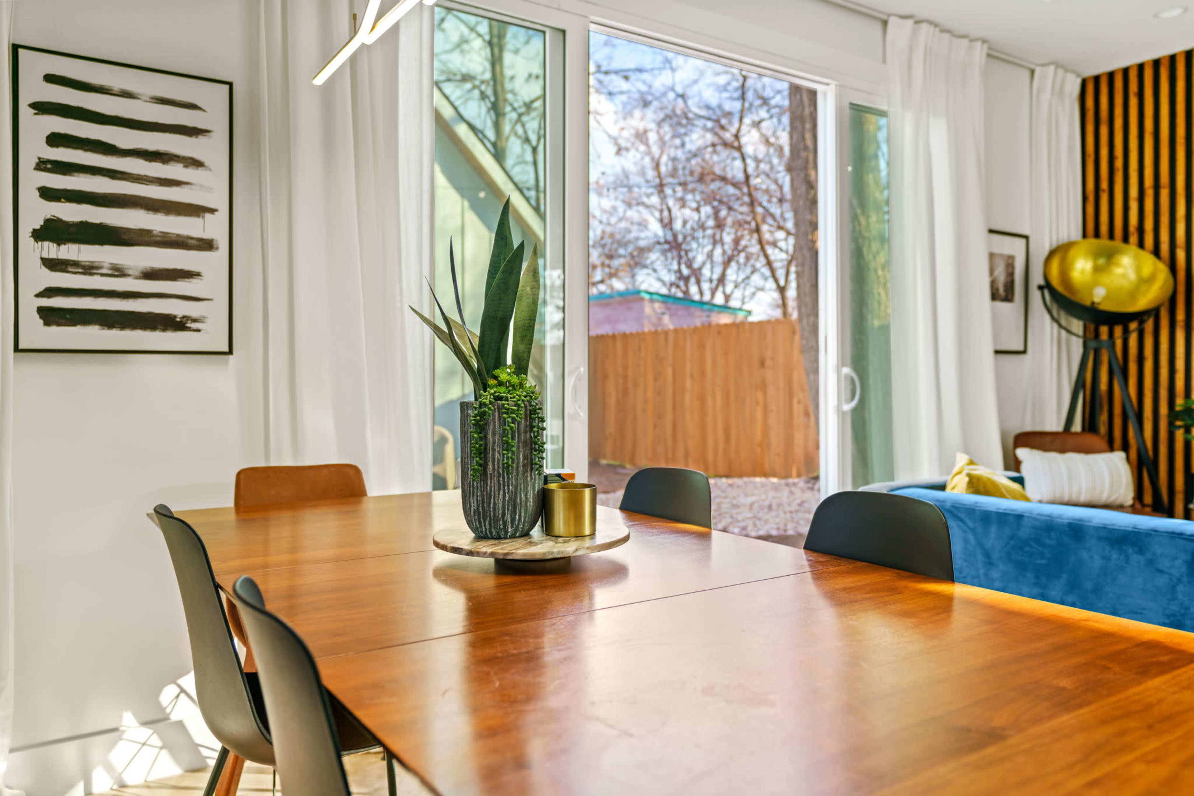 A wooden dining table is set with a plant centerpiece, surrounded by black chairs, with large windows revealing a backyard outside.