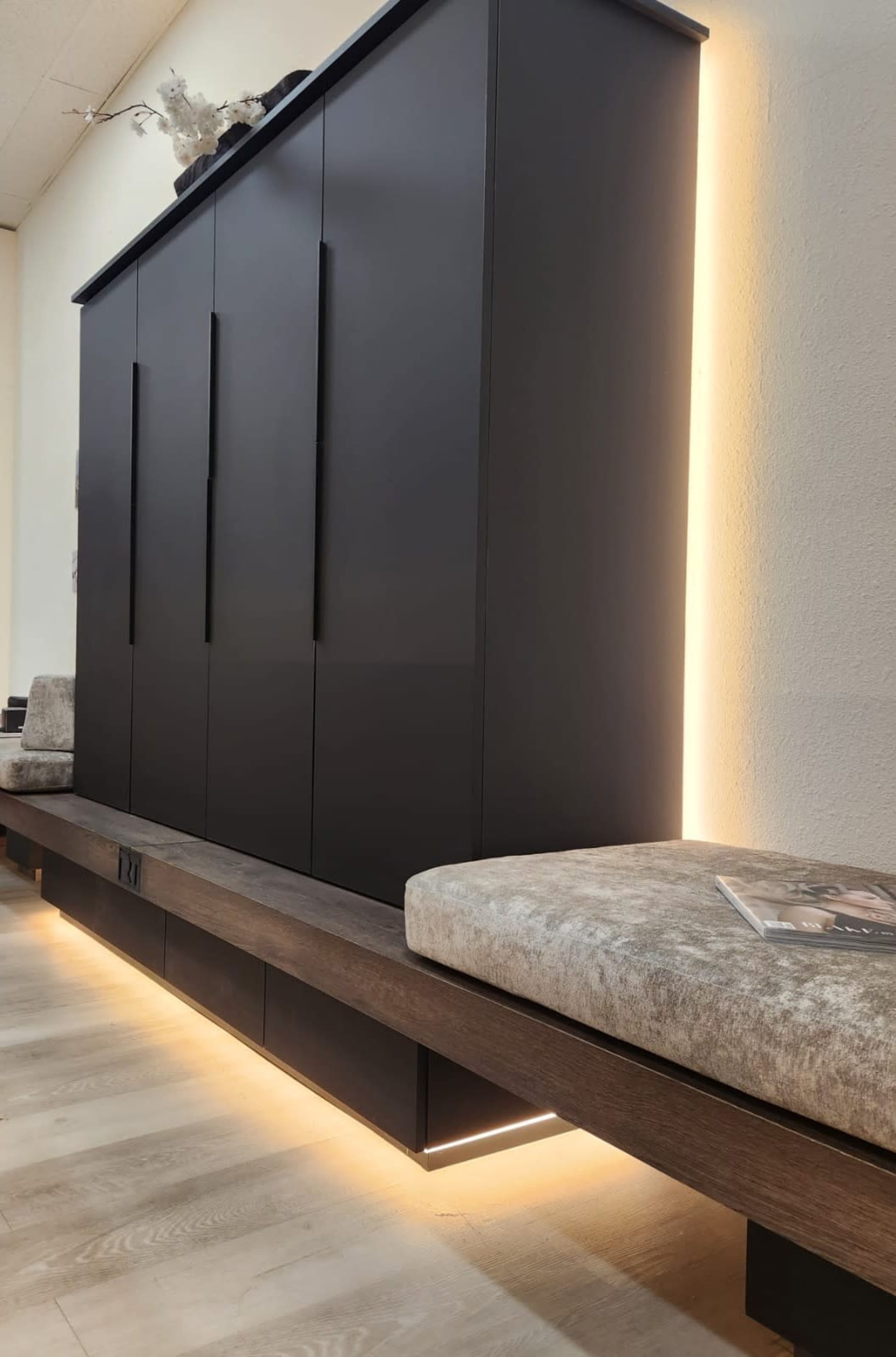 The image shows a modern, dark storage cabinet with sleek handles, accompanied by a low wooden bench featuring soft, upholstered seating and illuminated by a warm light strip along the bottom.