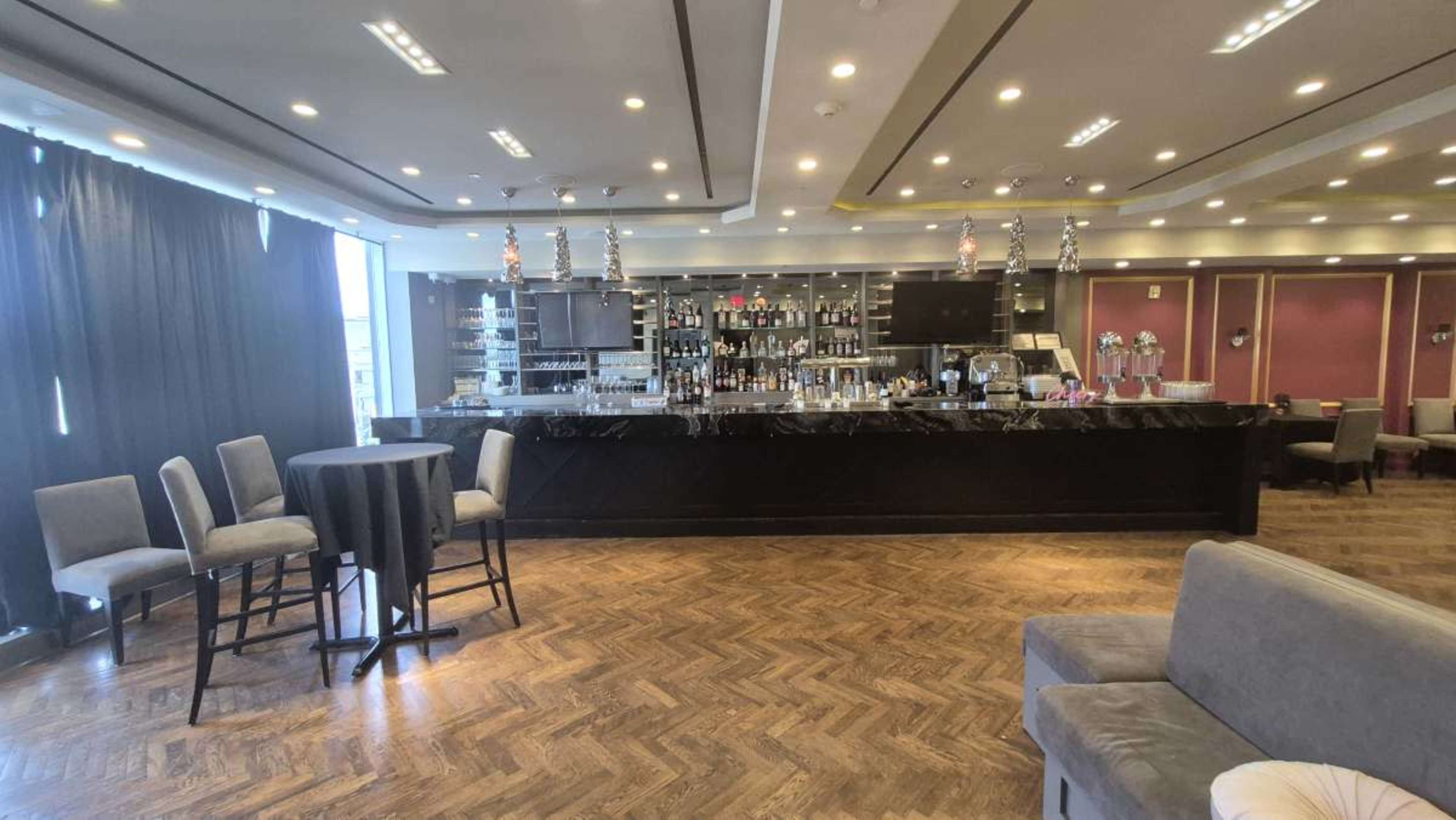 The image shows a modern bar interior with a sleek black bar counter, high chairs, and a well-stocked shelf of various bottles behind it, illuminated by overhead lights.