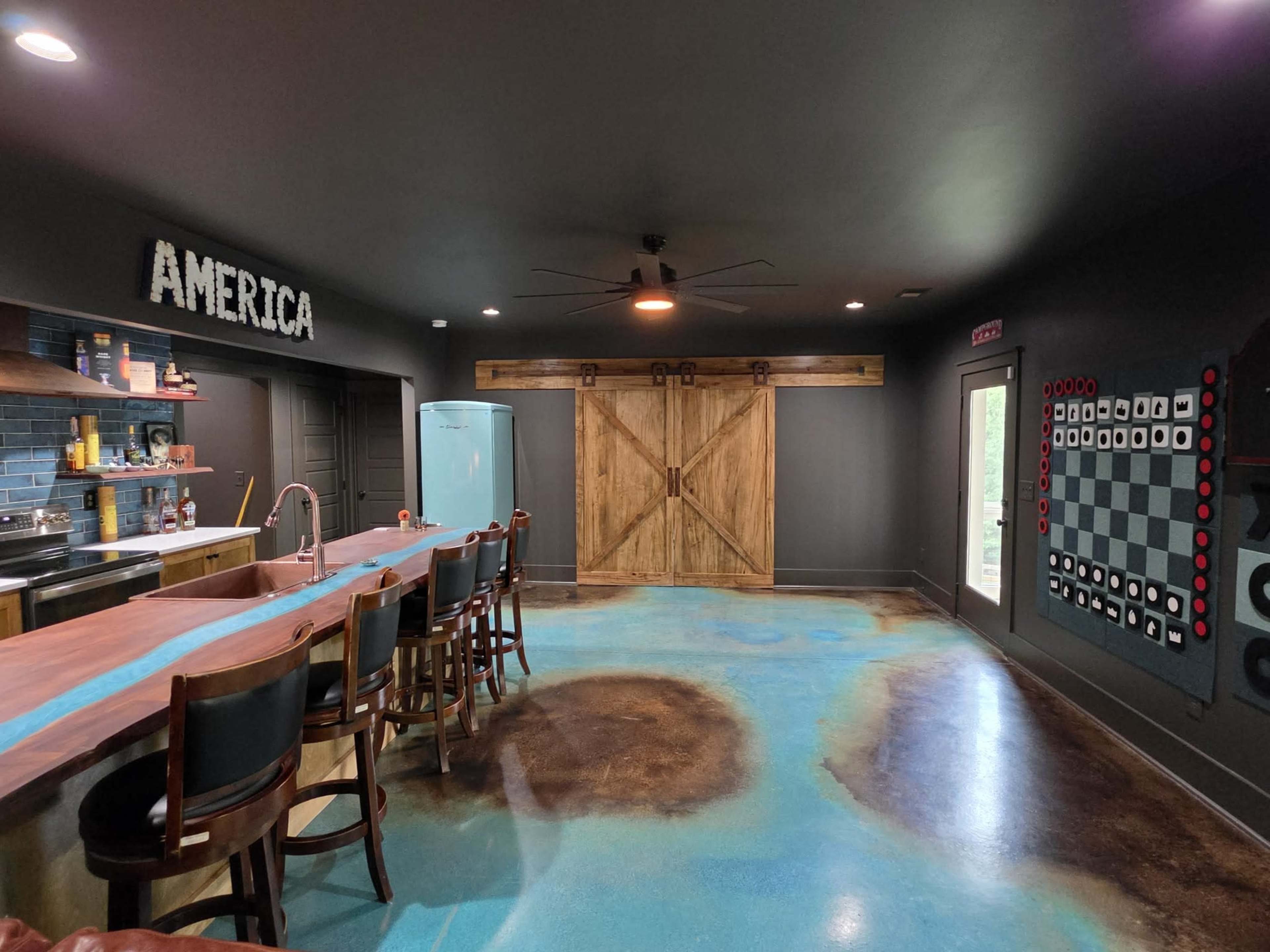 The image shows a room characterized by a bar with high stools, a kitchen area, and large wooden barn doors, with a floor featuring a unique color pattern.