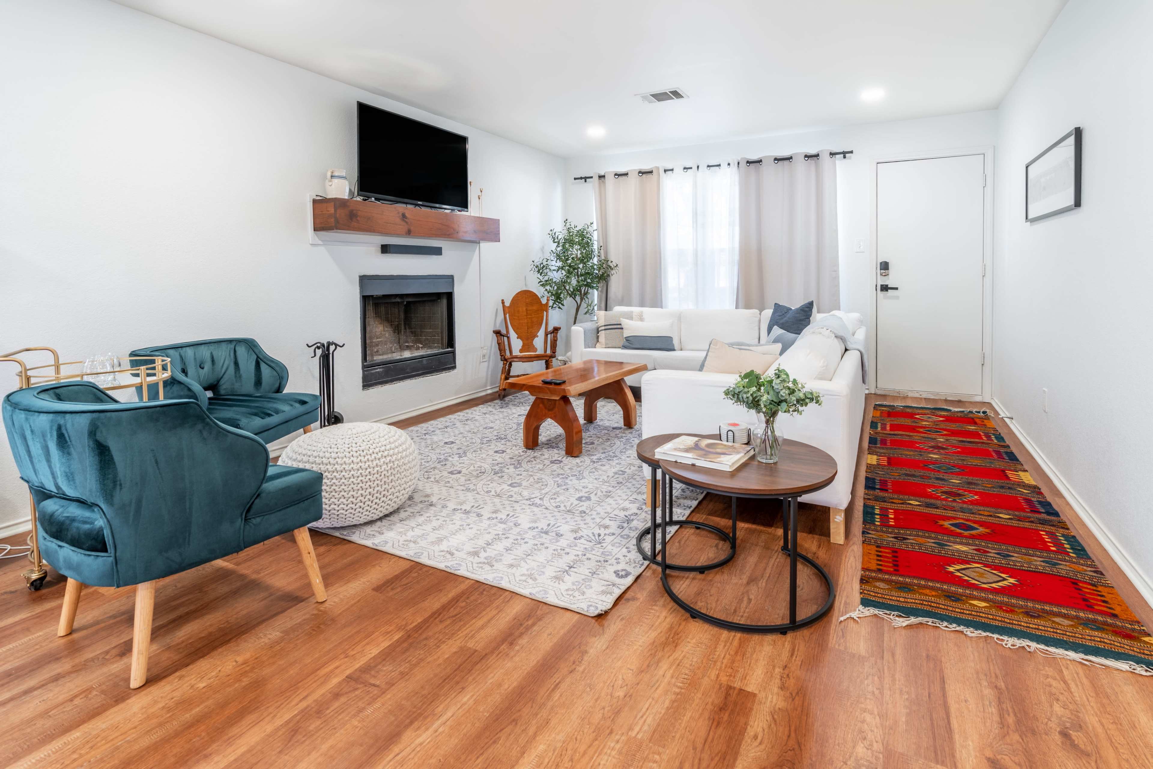 The living room features a cozy arrangement with a white sofa, wooden coffee table, and two teal chairs, set against a backdrop of a fireplace and a patterned area rug.