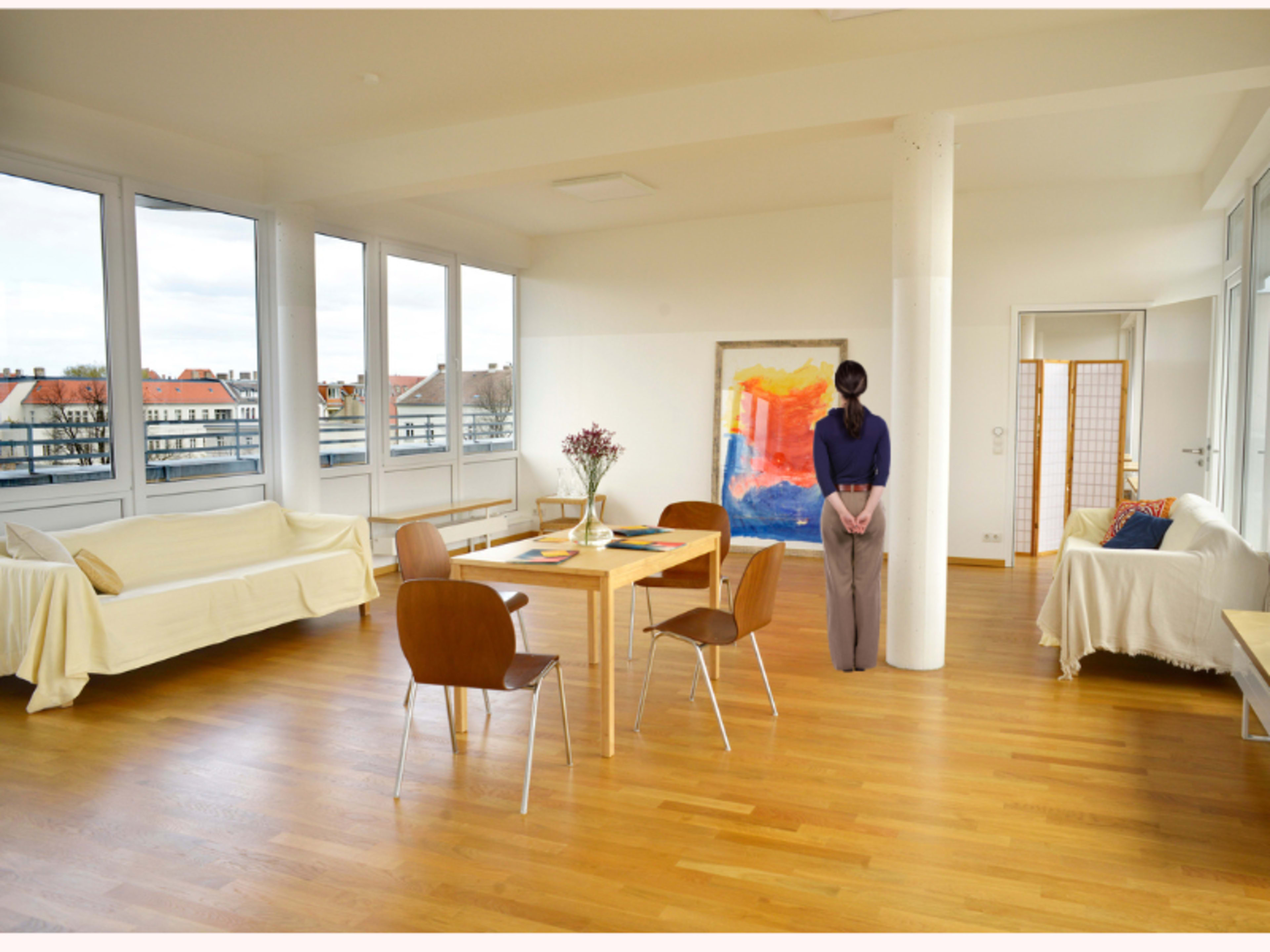 A spacious, modern living area features a dining table surrounded by chairs, a large artwork on the wall, and expansive windows overlooking a cityscape.