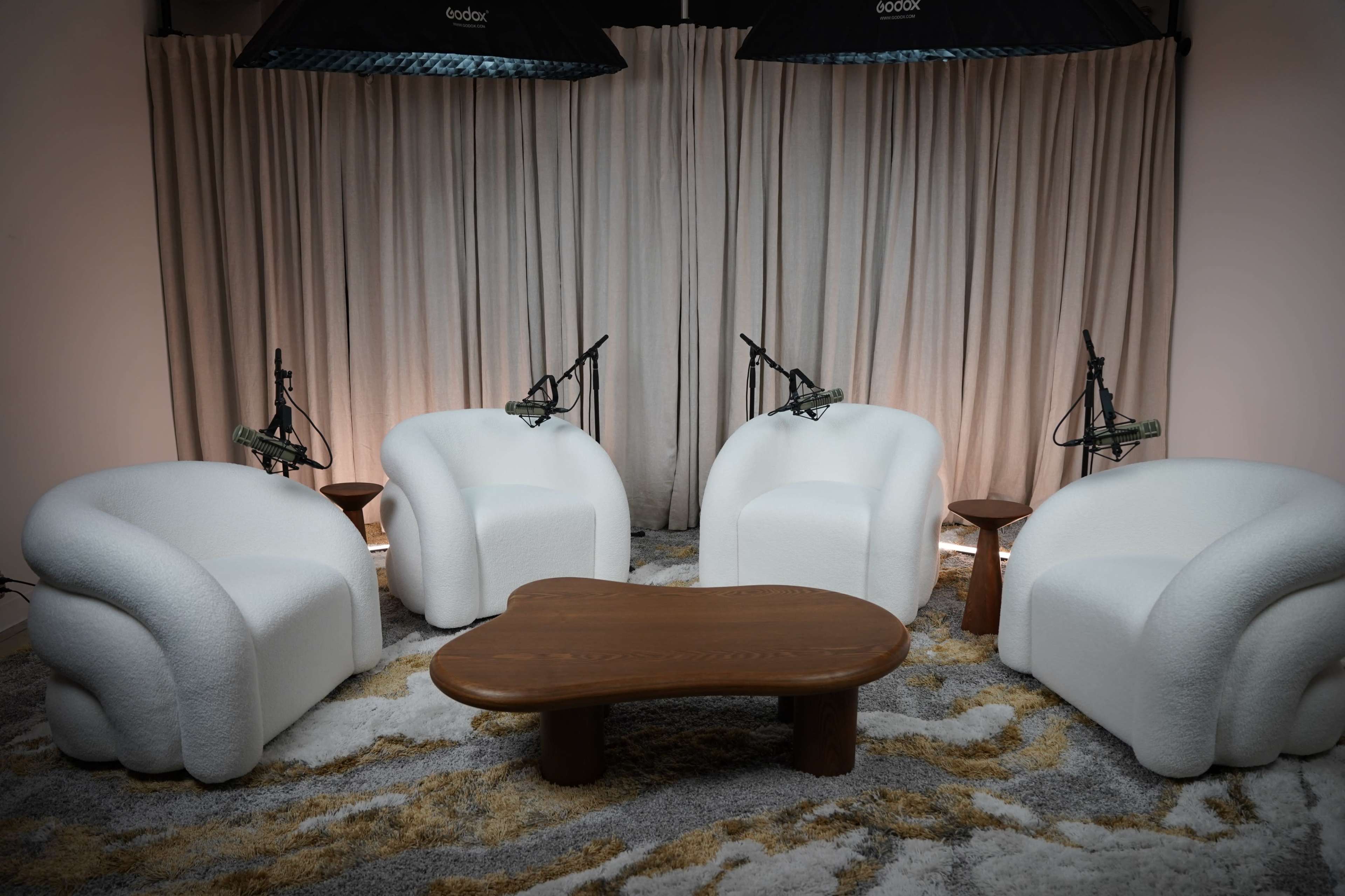 The image features a minimalist interior setup with four white, plush armchairs arranged around a wooden coffee table, under bright studio lighting and surrounded by microphone stands.