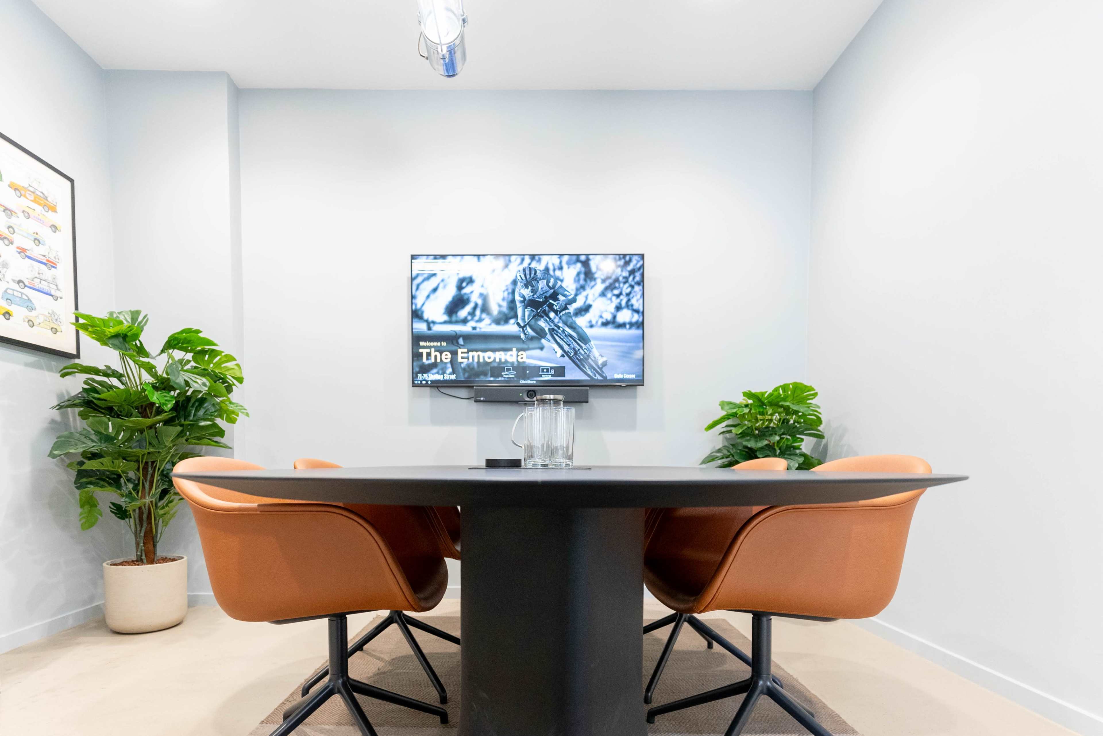 A conference room features a table with four brown chairs, a television mounted on the wall displaying a cycling image, and two potted plants in the corners.