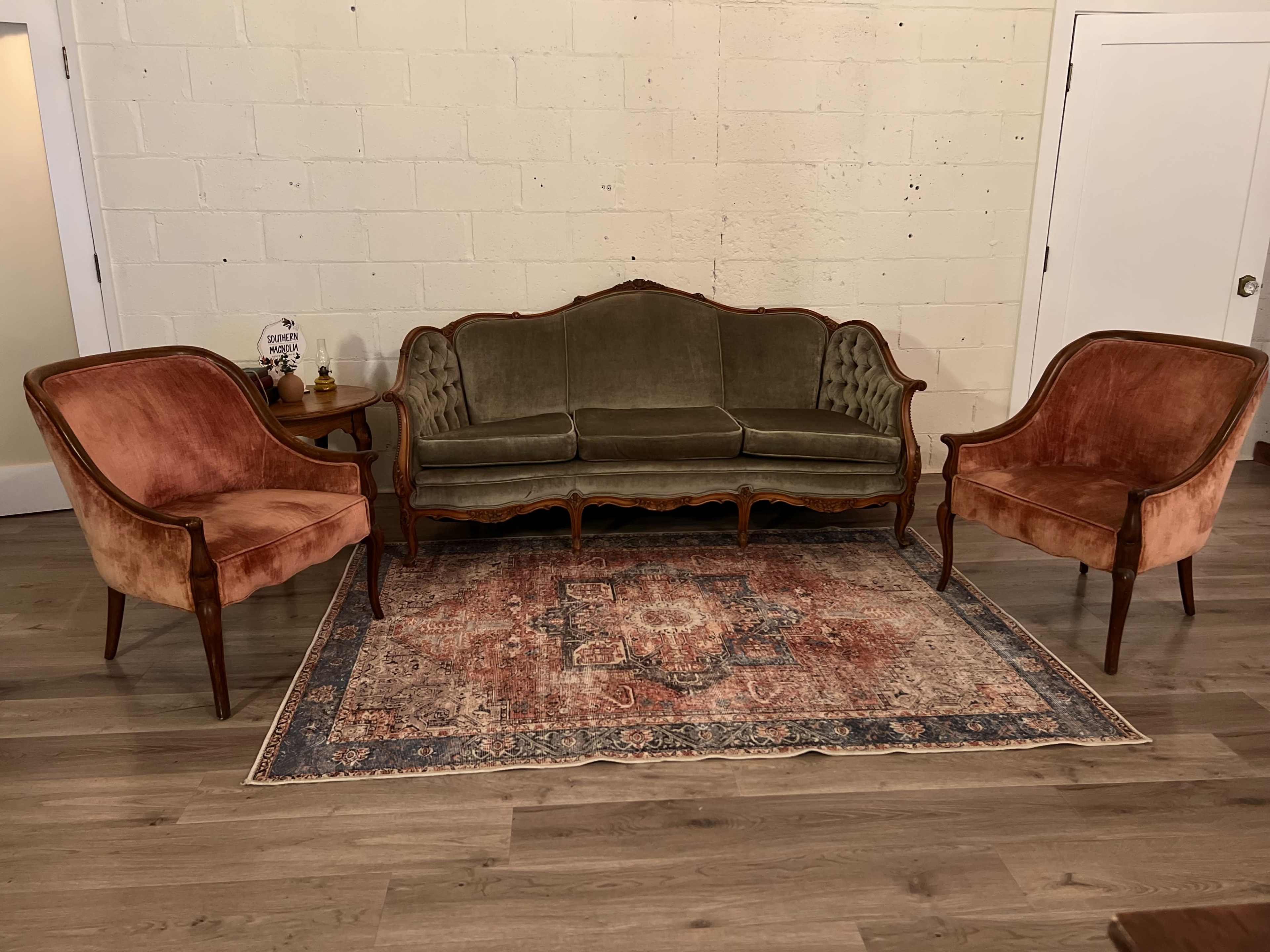 A vintage-style living room setup with a green tufted sofa and two pink upholstered chairs arranged around a patterned area rug.