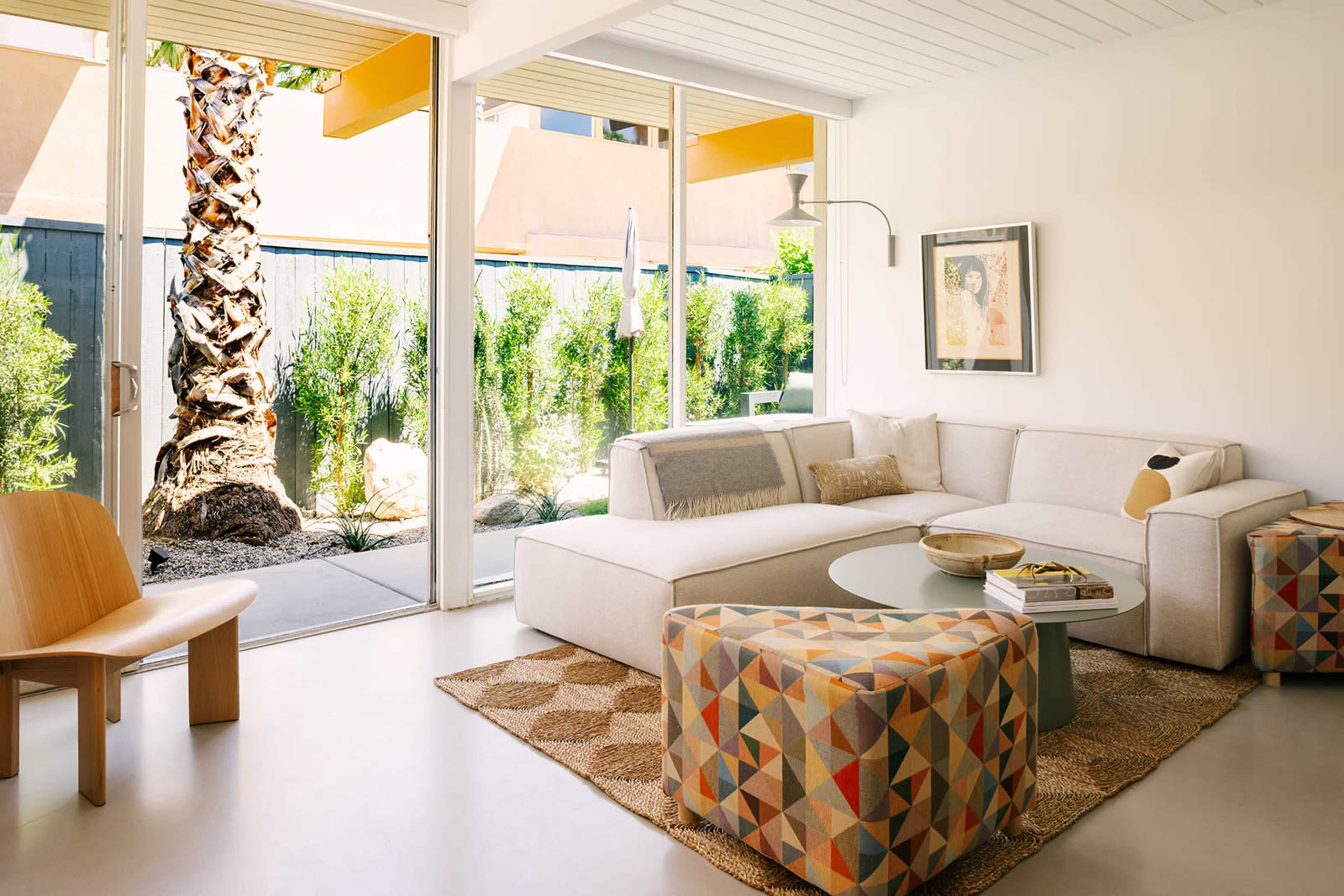 A modern living room features a sectional sofa, a round side table, and a colorful patterned ottoman, framed by large glass windows that overlook an outdoor space with greenery and a palm tree.