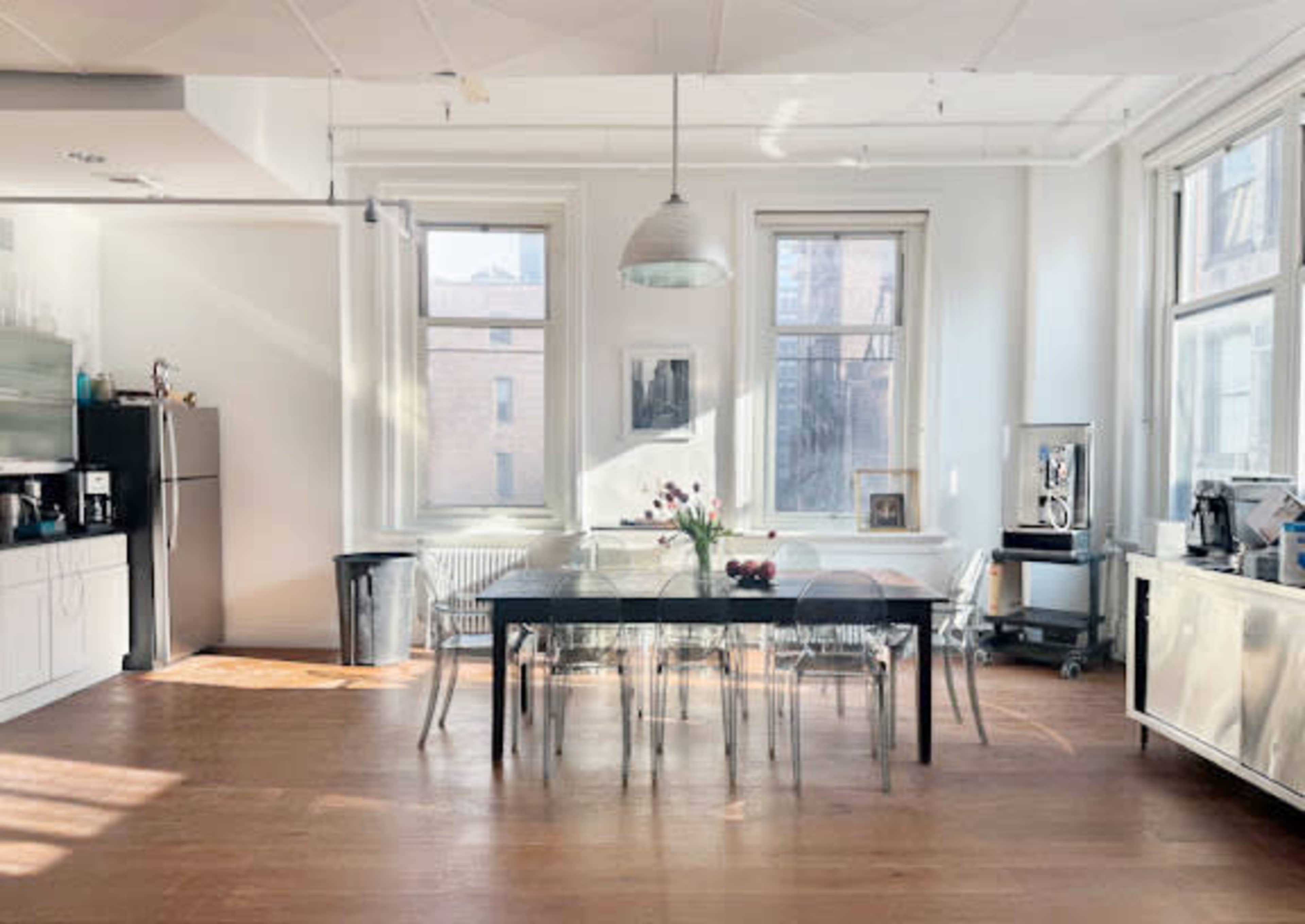 The image shows a bright, modern kitchen and dining area featuring a large black dining table surrounded by transparent chairs, large windows allowing natural light in, and kitchen appliances visible on one side.