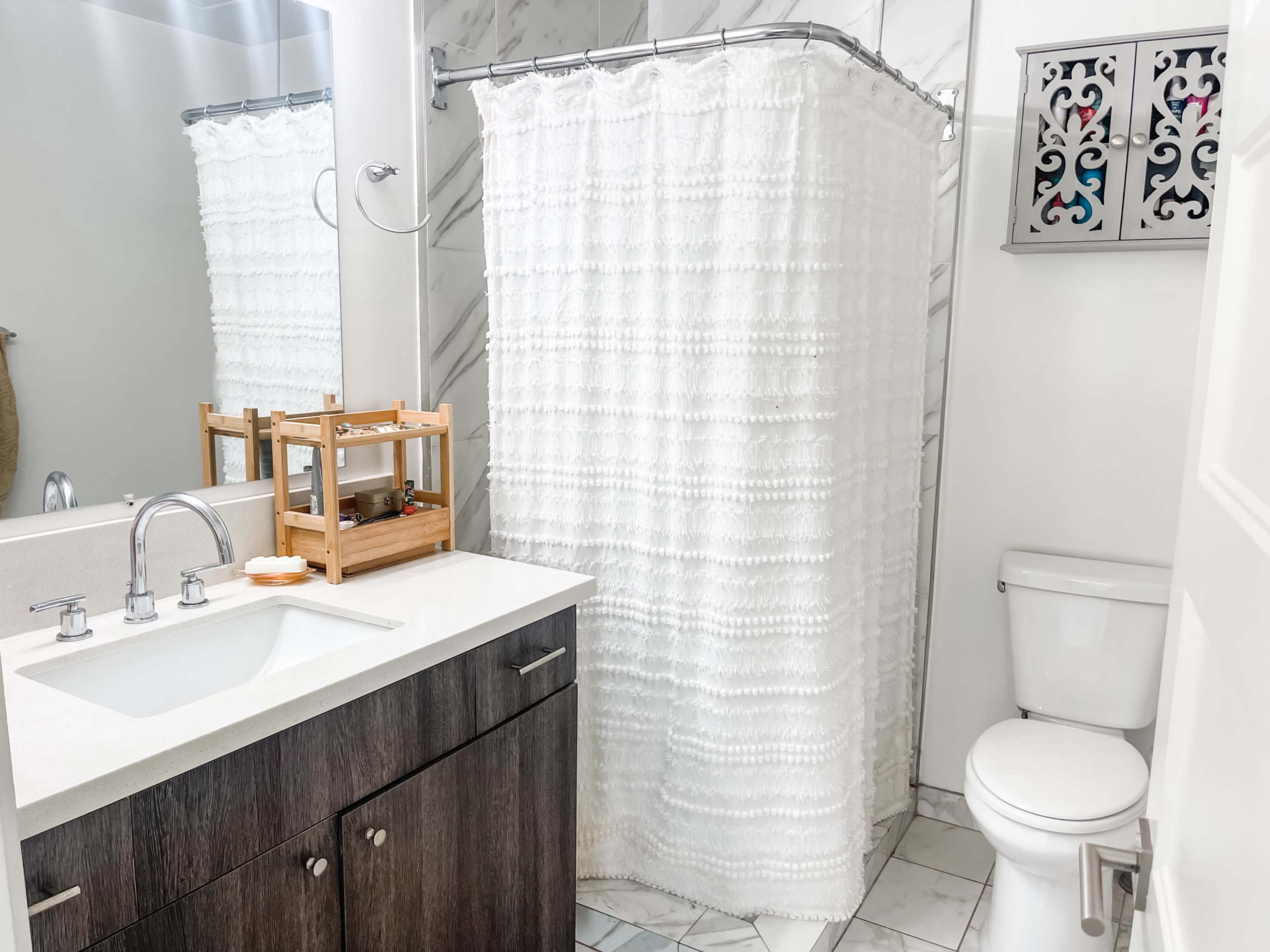 A bathroom features a white shower curtain, a wooden storage rack, a sink with a stone countertop, and a toilet against tiled walls.