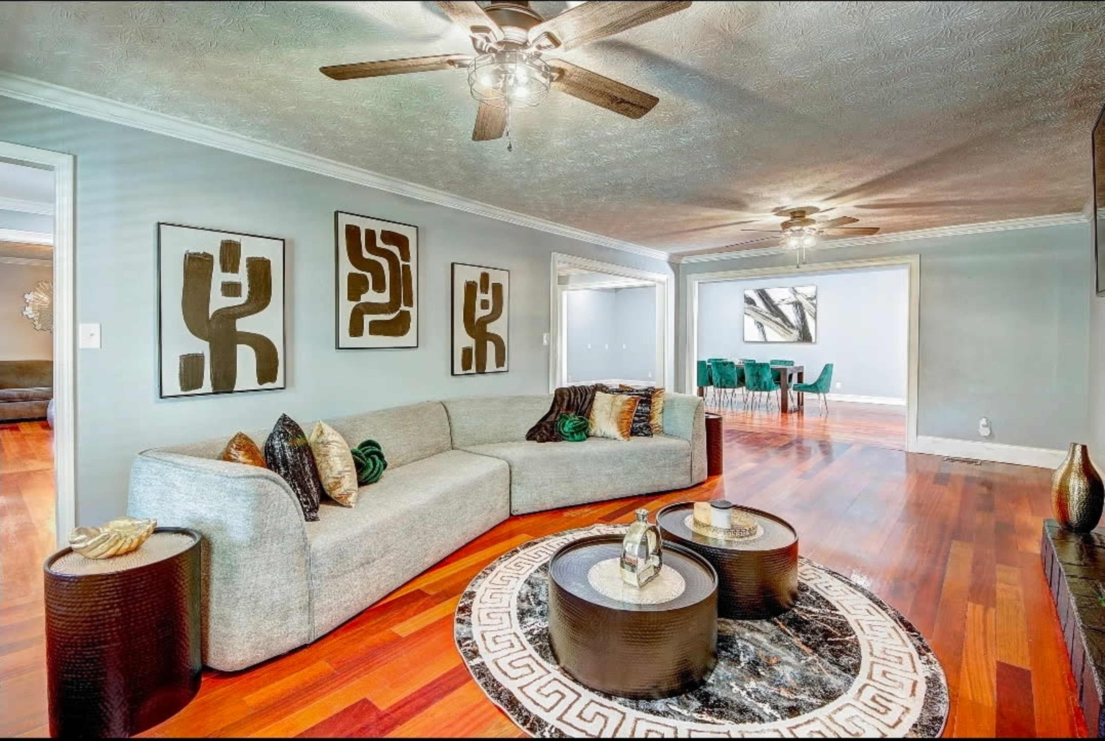 The living room features a light gray sectional sofa, a round area rug, and abstract wall art, with a view into a dining area in the background.
