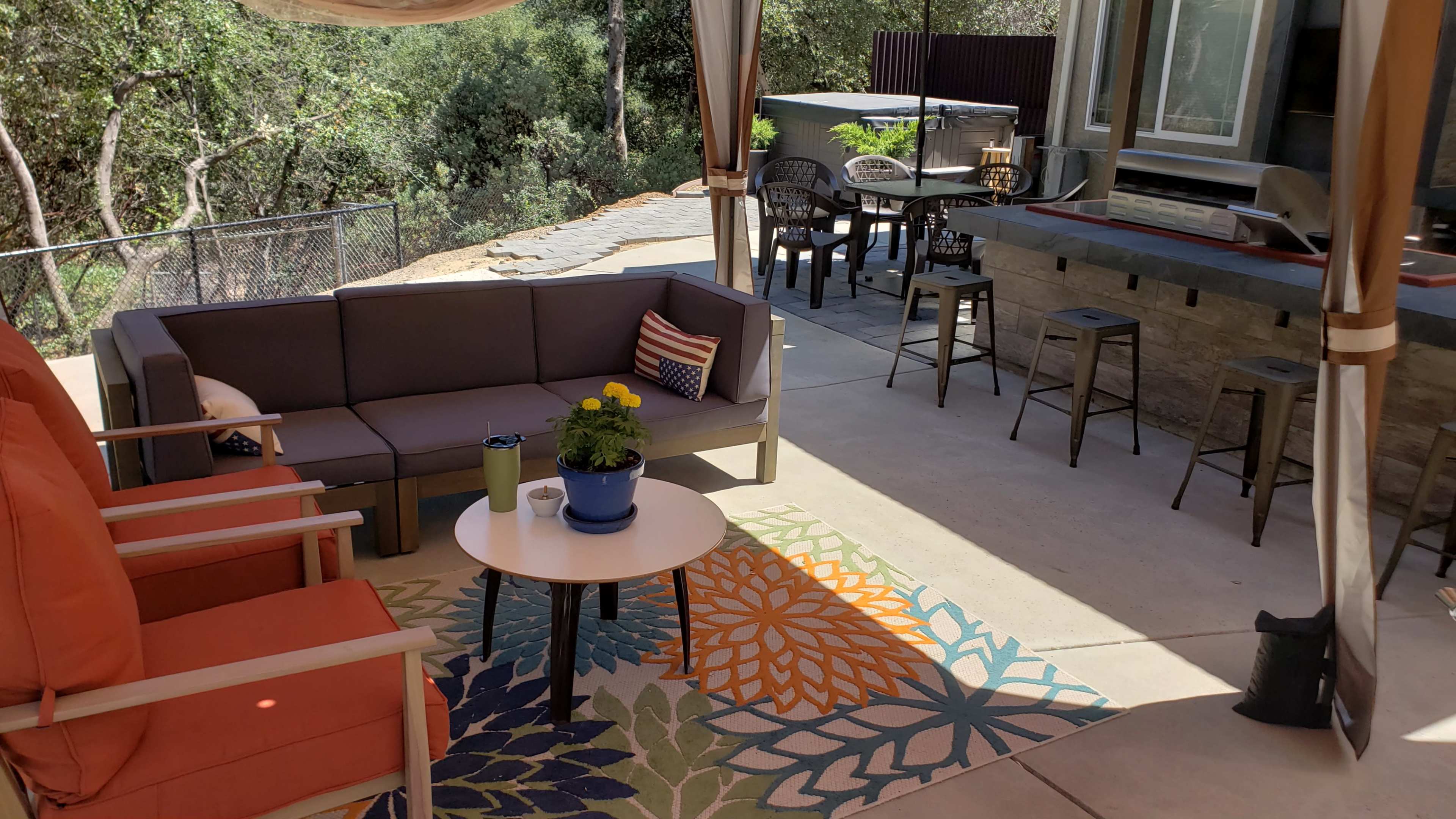 The scene shows a covered outdoor seating area with a sofa, armchairs, a table, and a colorful rug, alongside a dining set and modern bar in a garden setting.