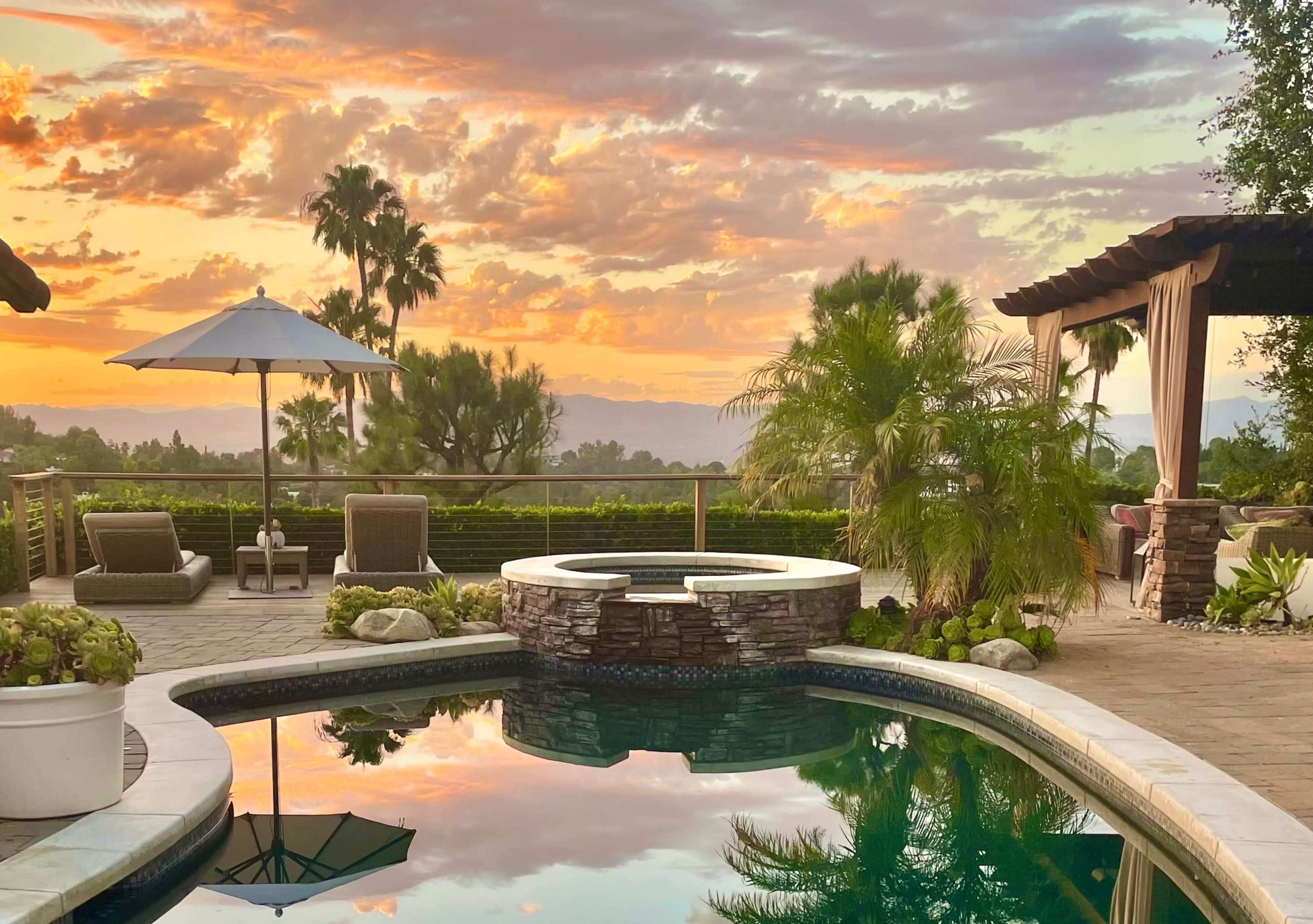 The scene features a tranquil backyard with a swimming pool reflecting a vibrant sunset, surrounded by palm trees and lounge chairs.