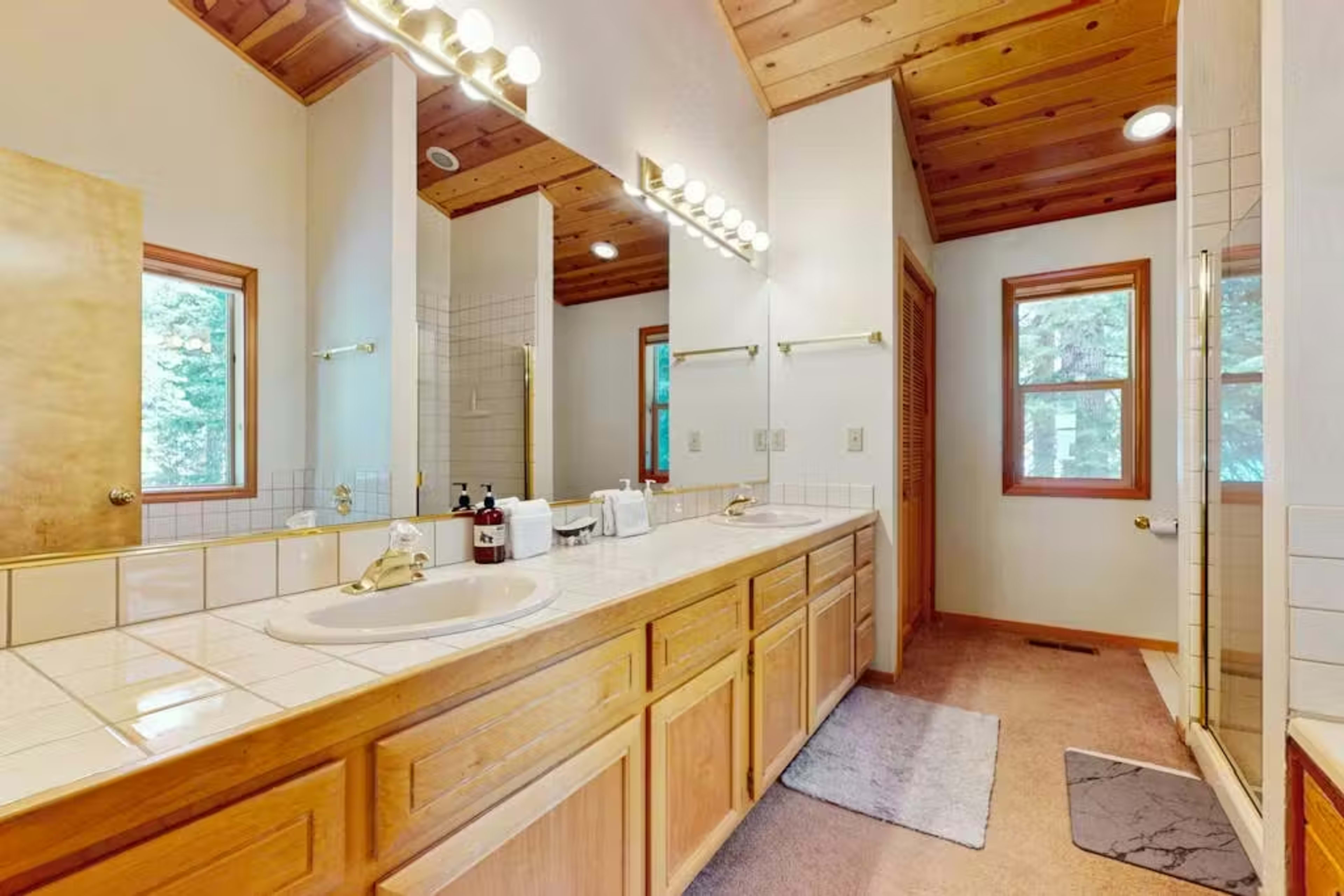 The bathroom features a double vanity with wooden cabinets, light-colored tile countertops, and large windows that provide natural light.