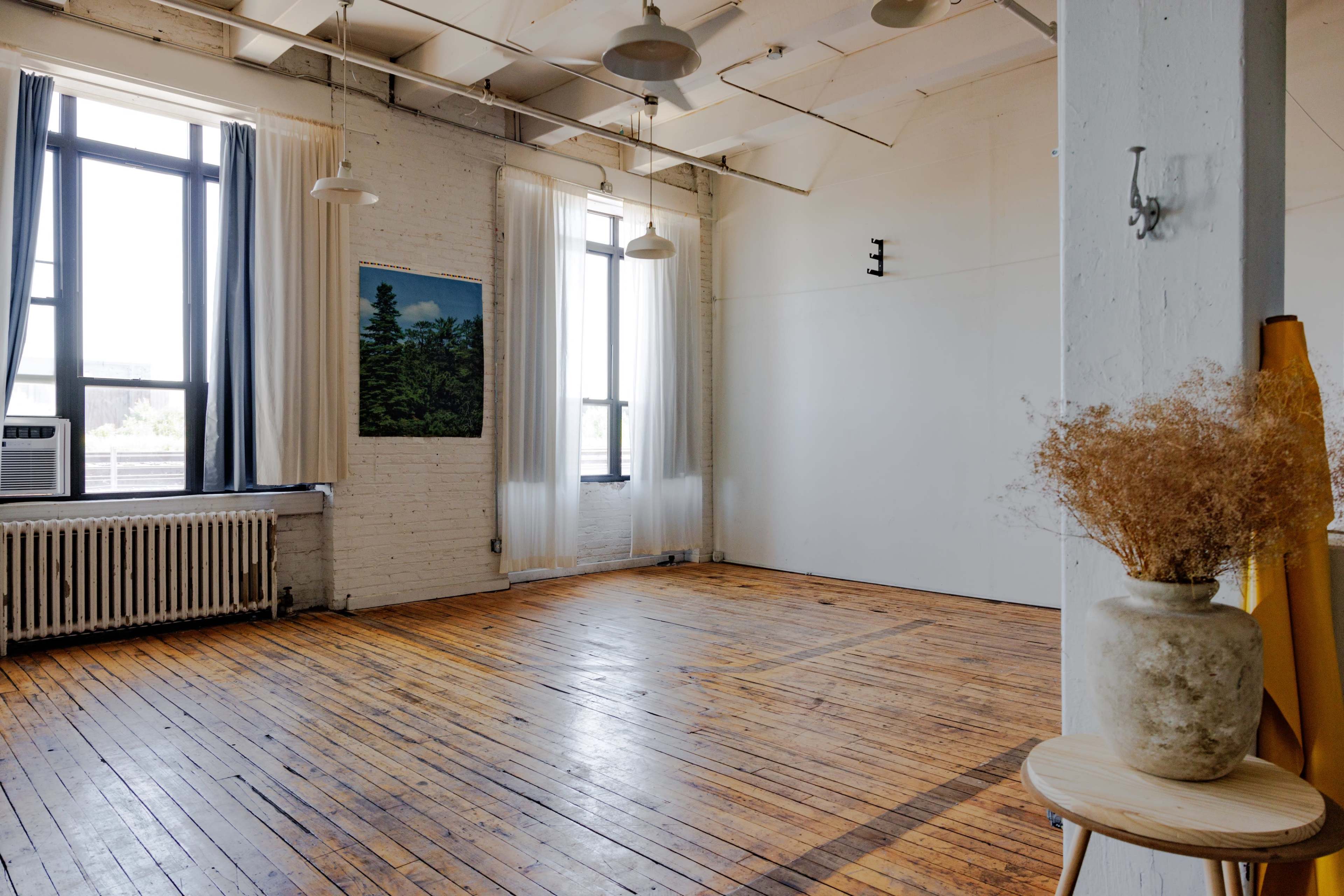 A light-filled, spacious room with wooden floors, large windows, and minimal furnishings, including a plant arrangement on a small table.