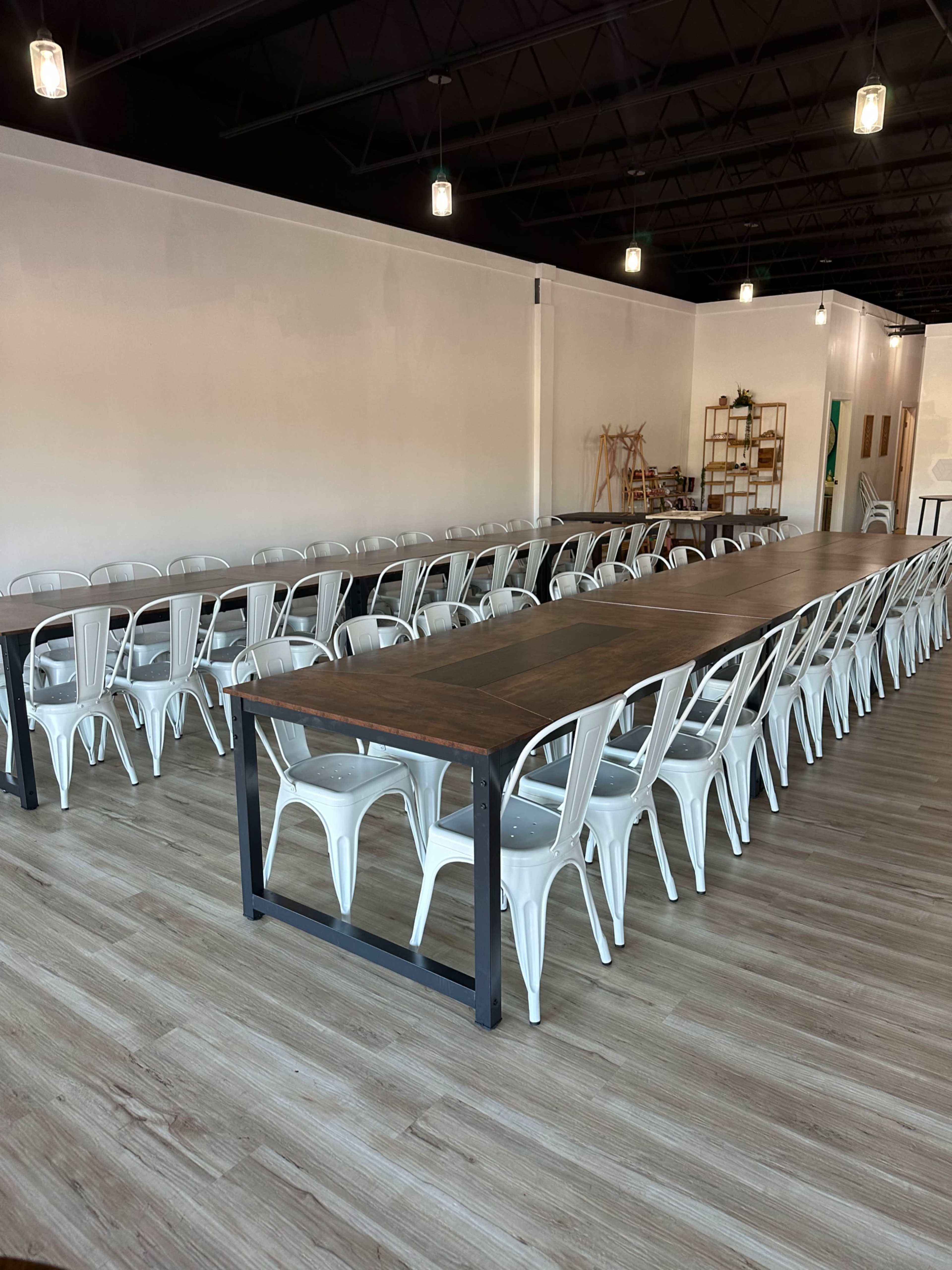 A long wooden table is arranged with white metal chairs in a spacious, well-lit room with wooden flooring.