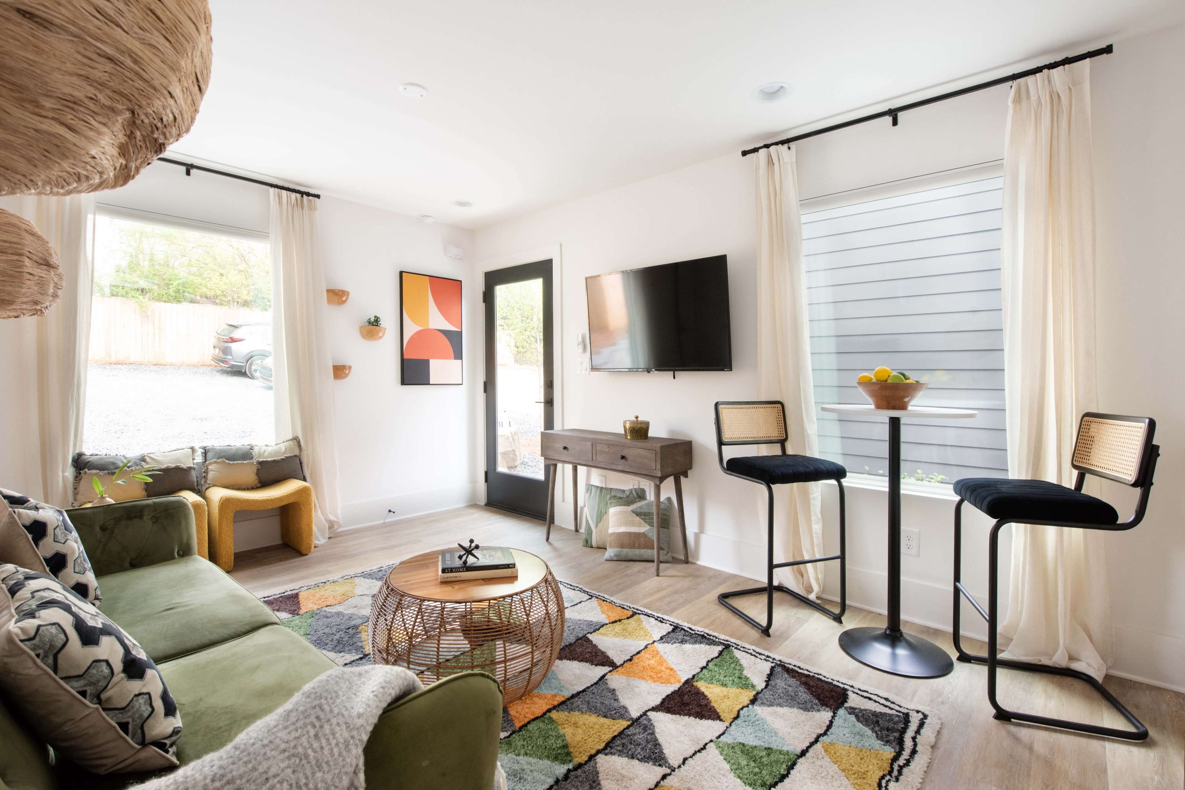The image shows a modern living room with a green sofa, a round coffee table on a colorful geometric rug, and two bar stools near a console table, adjacent to large windows and a door leading outside.