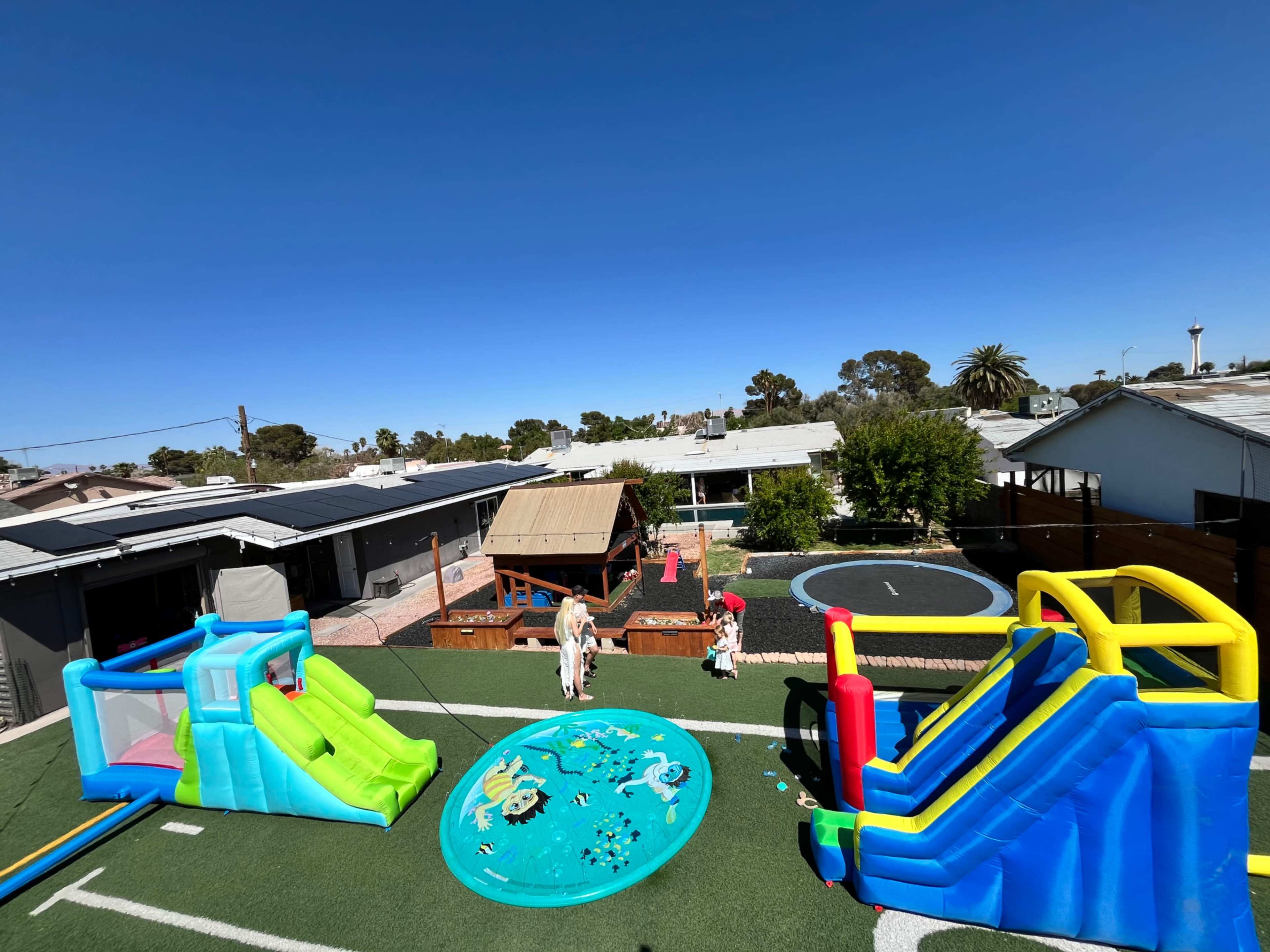 The image shows a backyard playground featuring inflatable slides, a sandbox, and a trampoline under a clear blue sky.