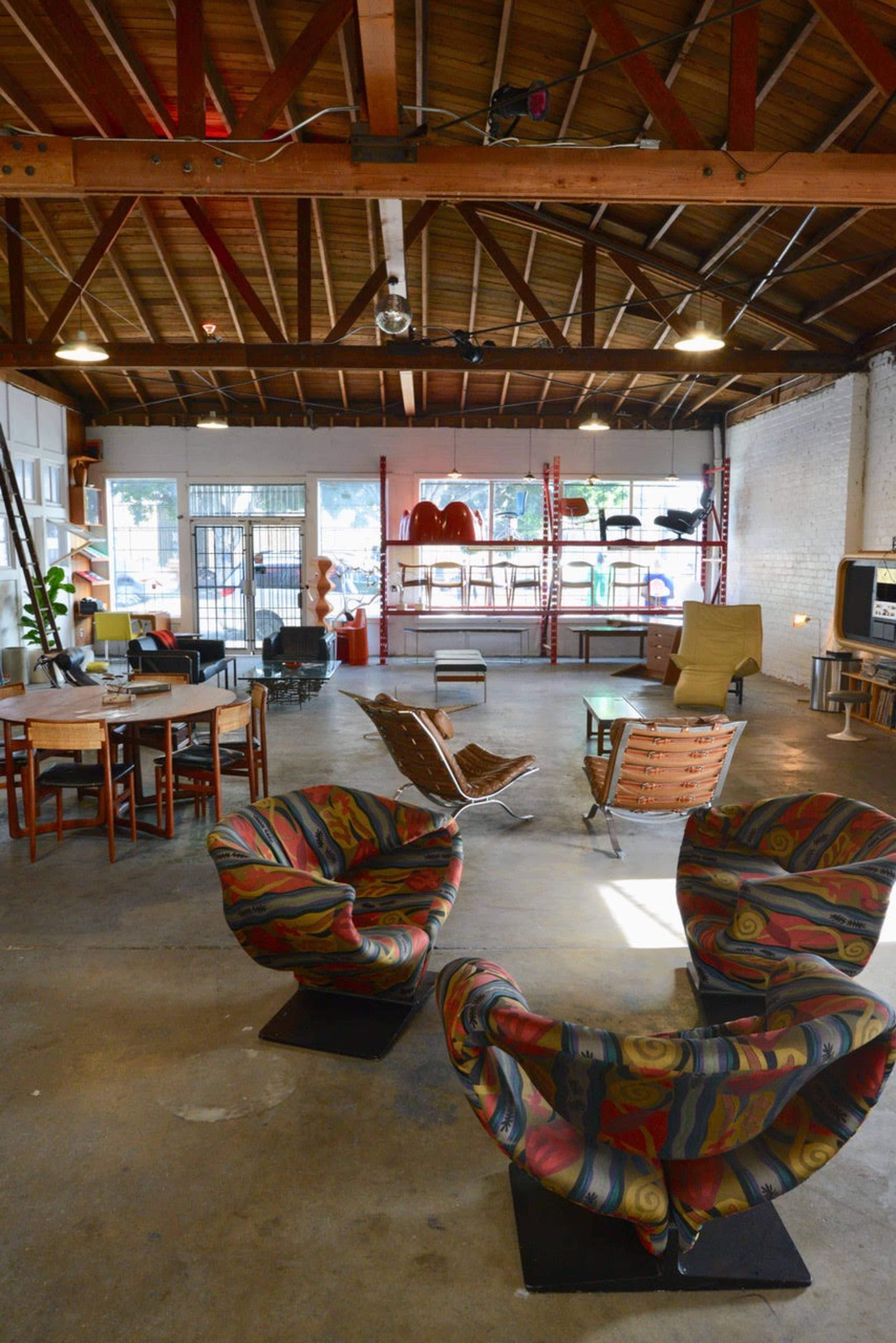 An open, industrial-style interior with various seating arrangements, including colorful chairs, a dining table, and shelving units in the background.