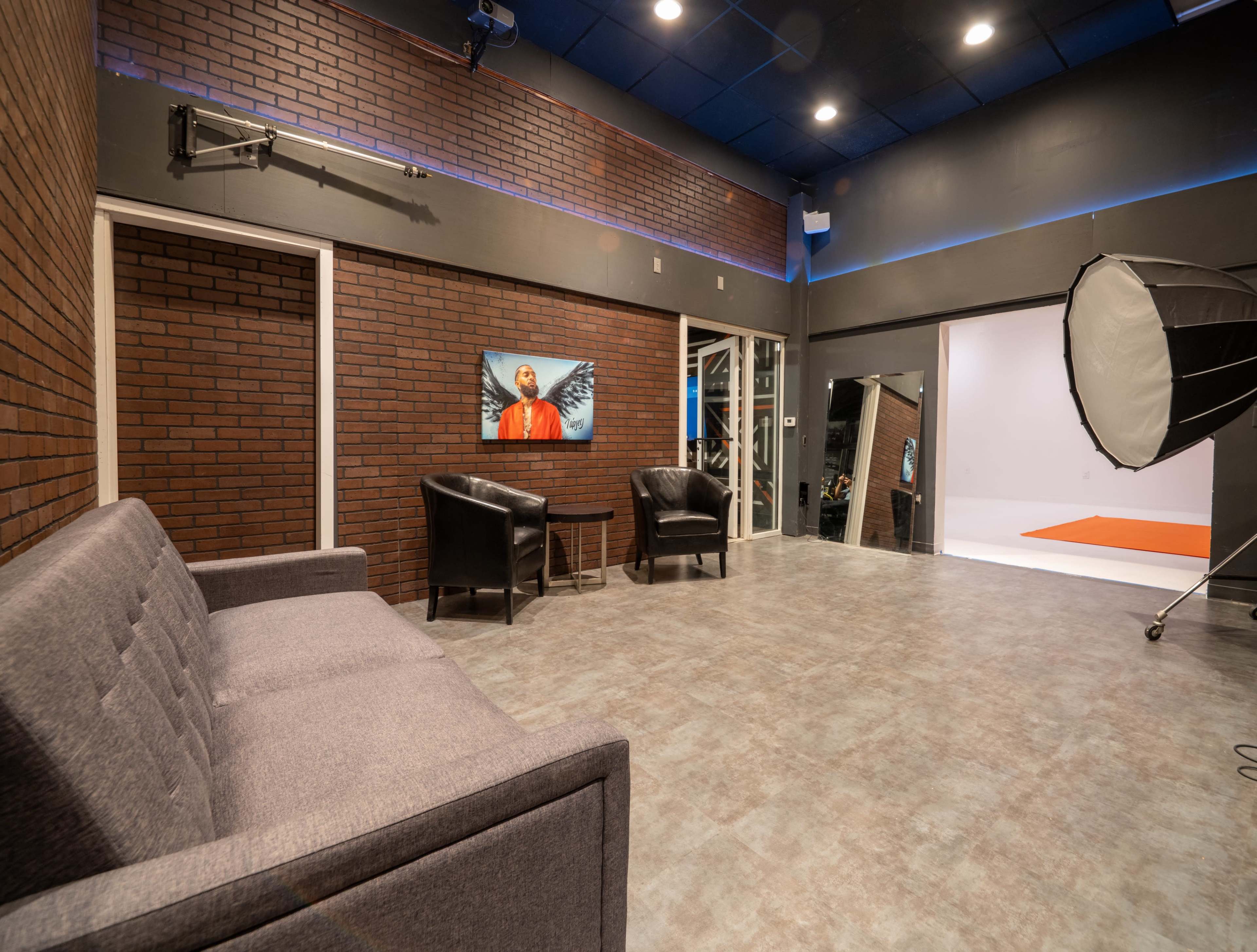 A photography studio features a gray couch, two black chairs, and a large backdrop area with bright lighting.