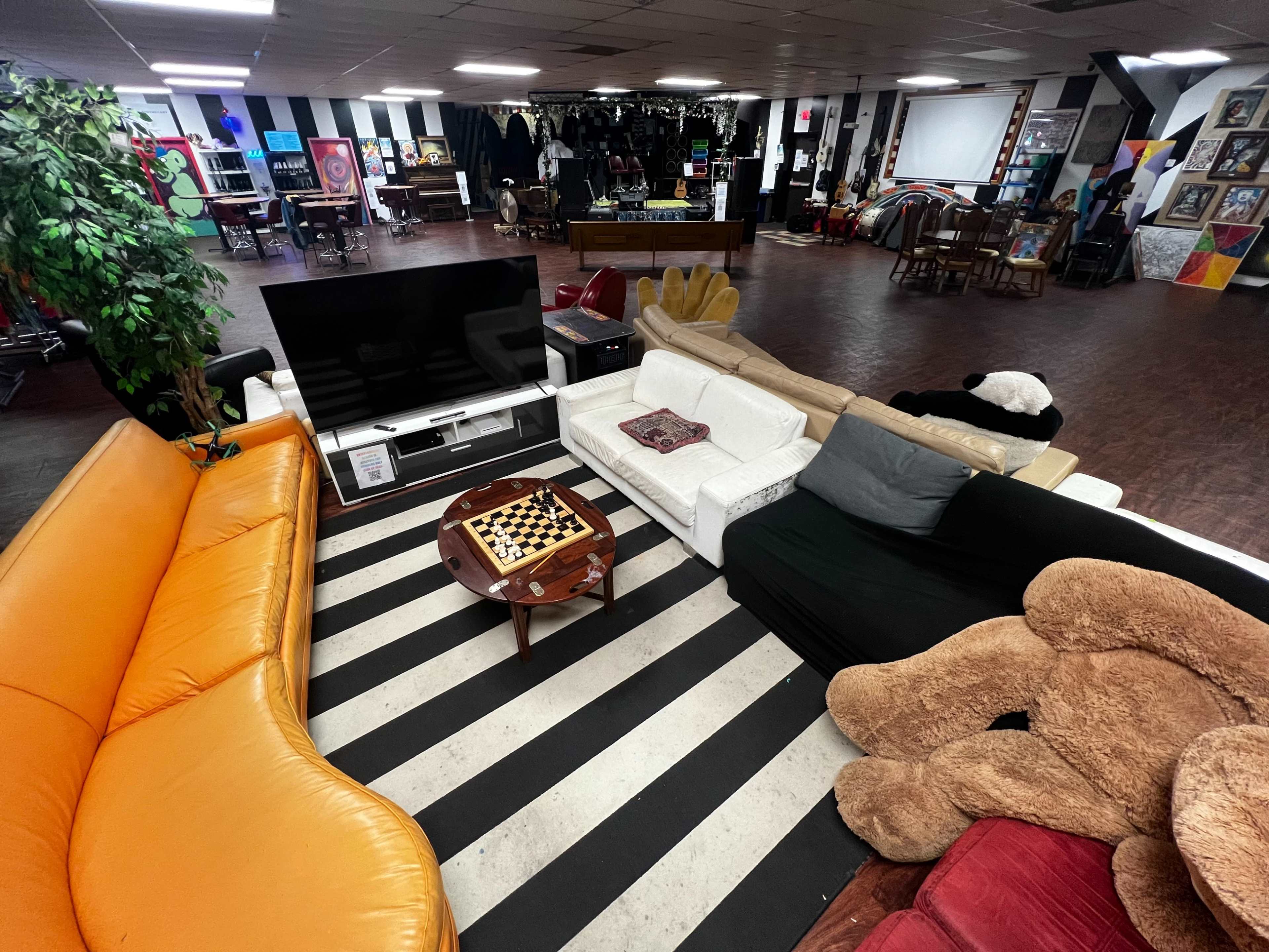 The image shows a large, open lounge area with various couches arranged around a coffee table on a striped rug, featuring a television and diverse artwork in the background.