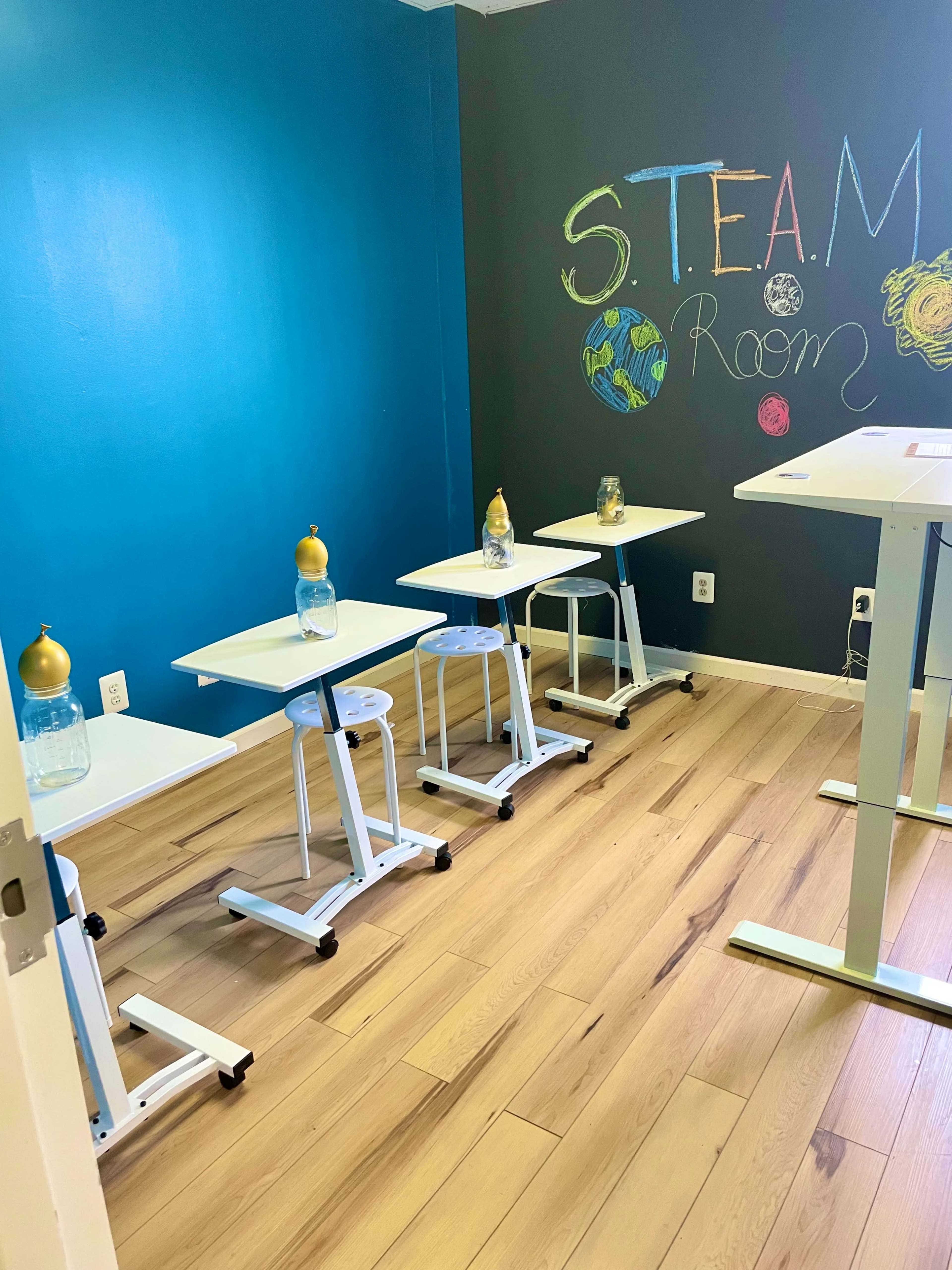 A small classroom with four movable desks, each featuring a water bottle, and a wall painted with "S.T.E.A.M. Room."