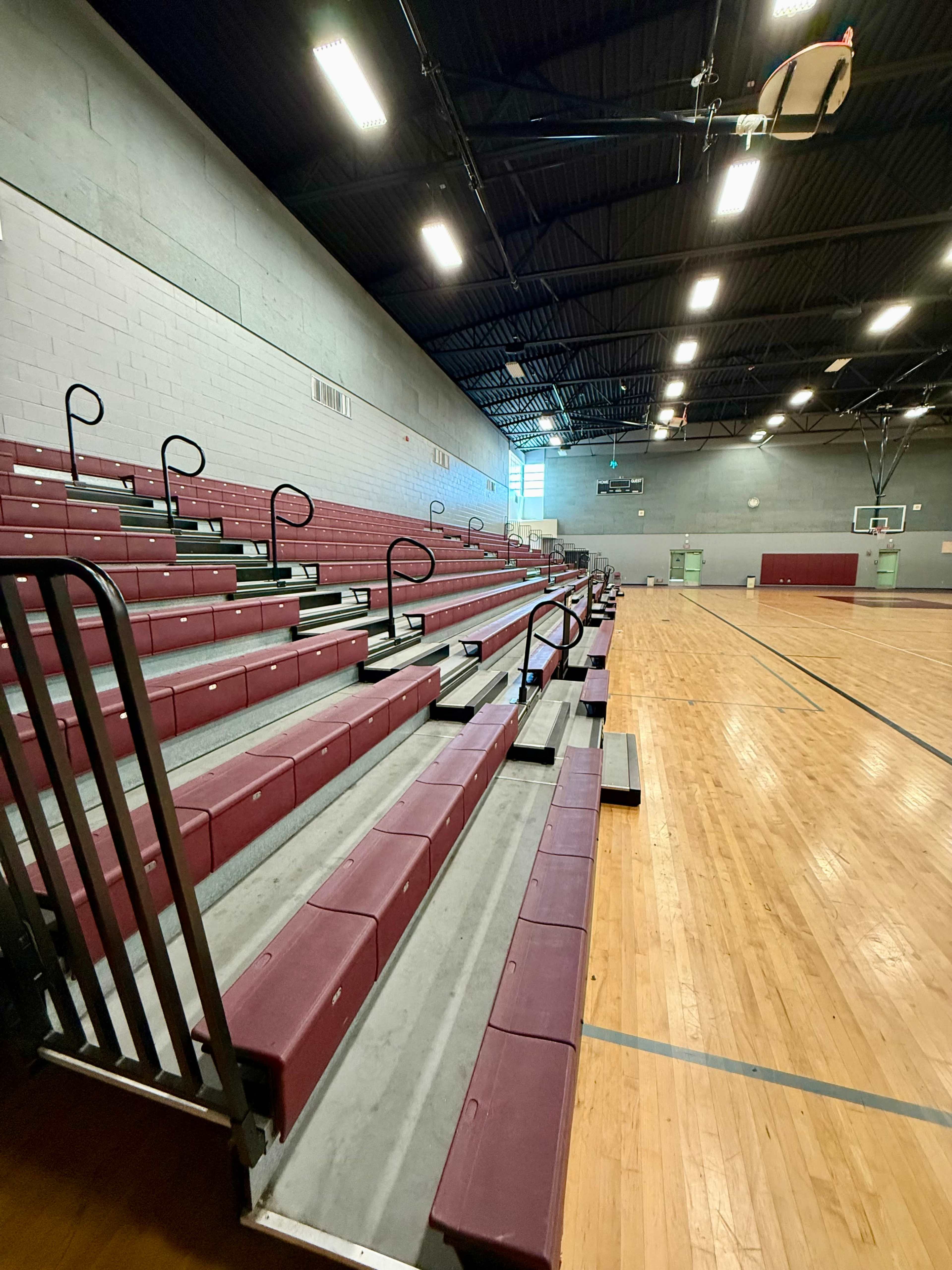 The image shows empty bleacher seating in a gymnasium with wooden floors and basketball hoops in the background.