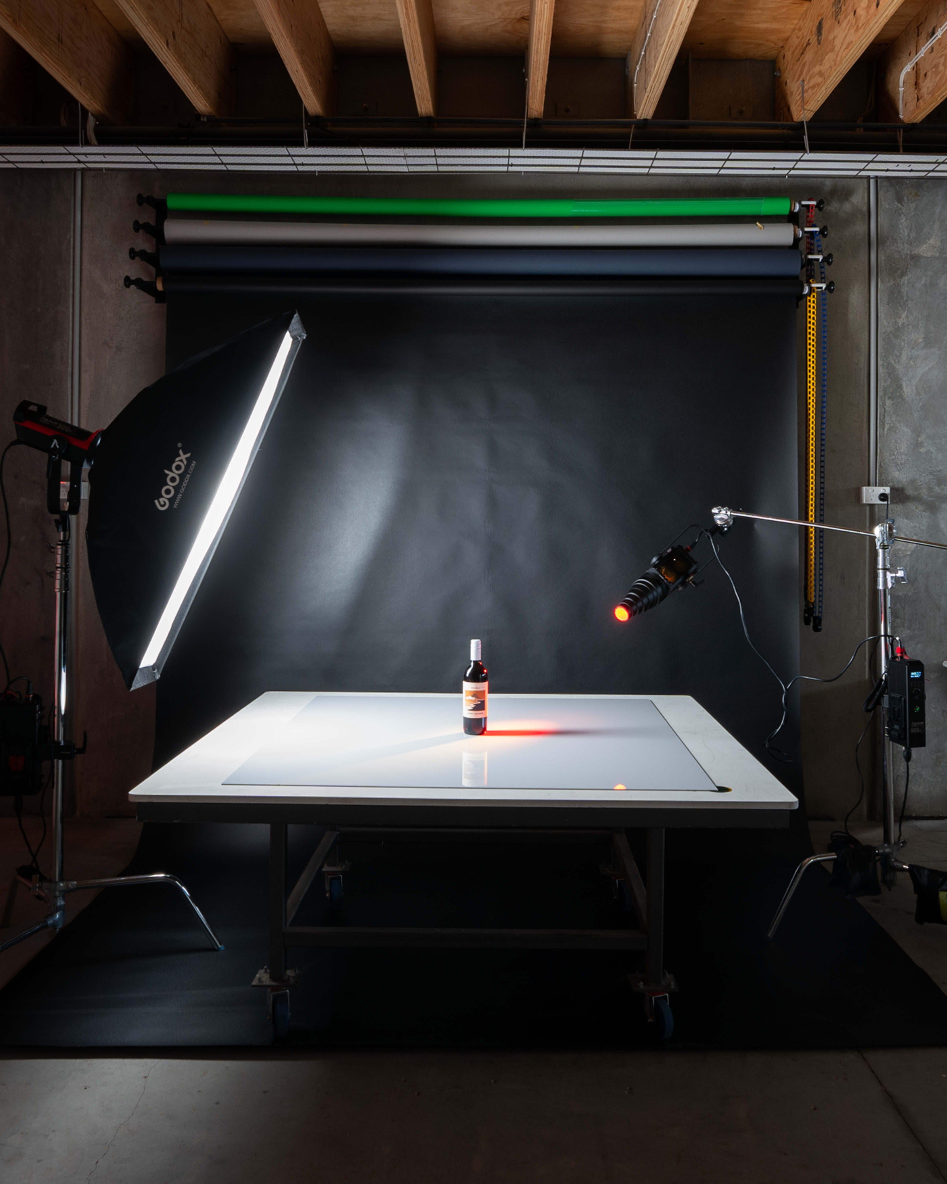 A wine bottle is placed on a light table, surrounded by photography lighting equipment and a black backdrop.