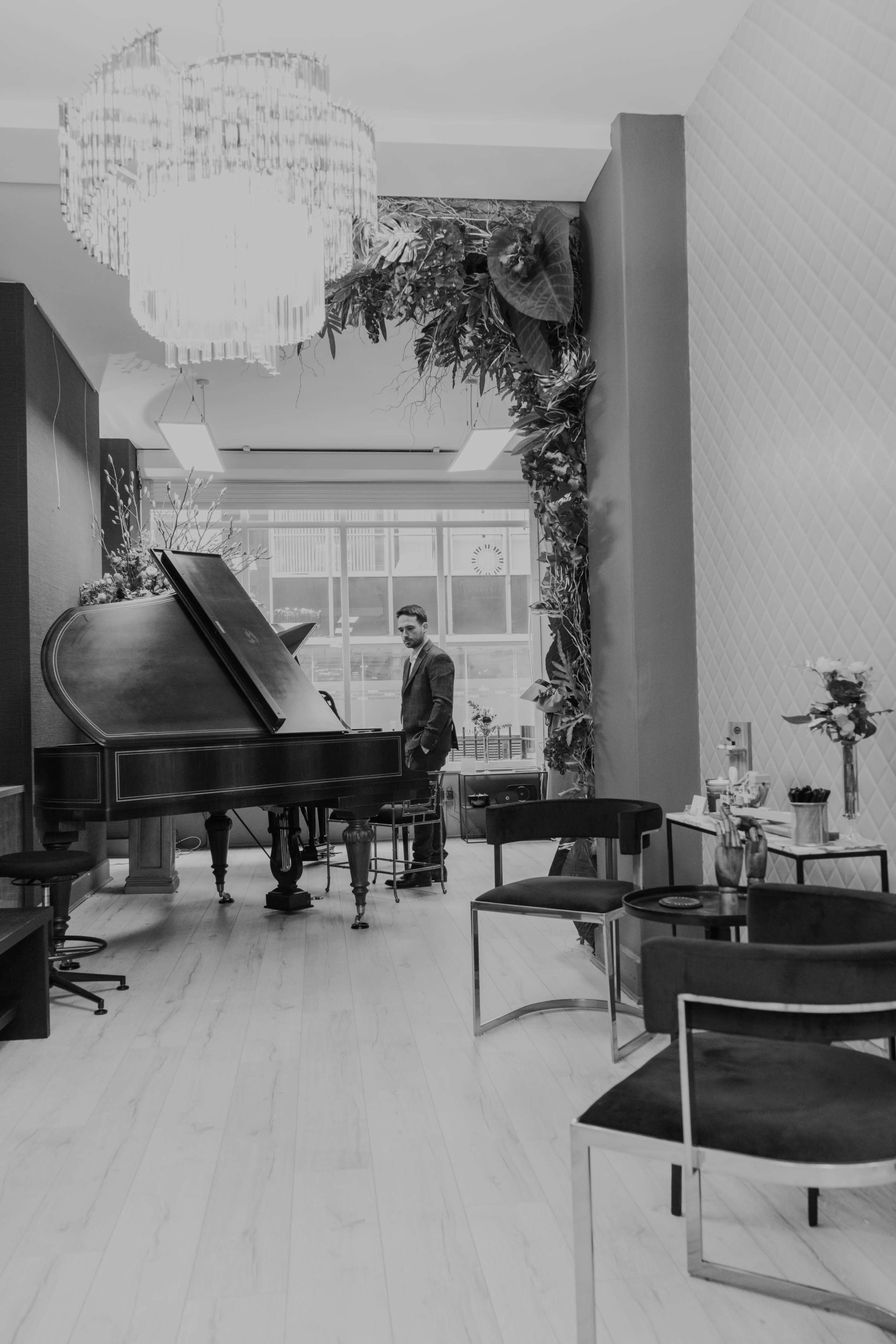A man stands beside a grand piano in a modern, plant-decorated room with stylish seating and a chandelier.