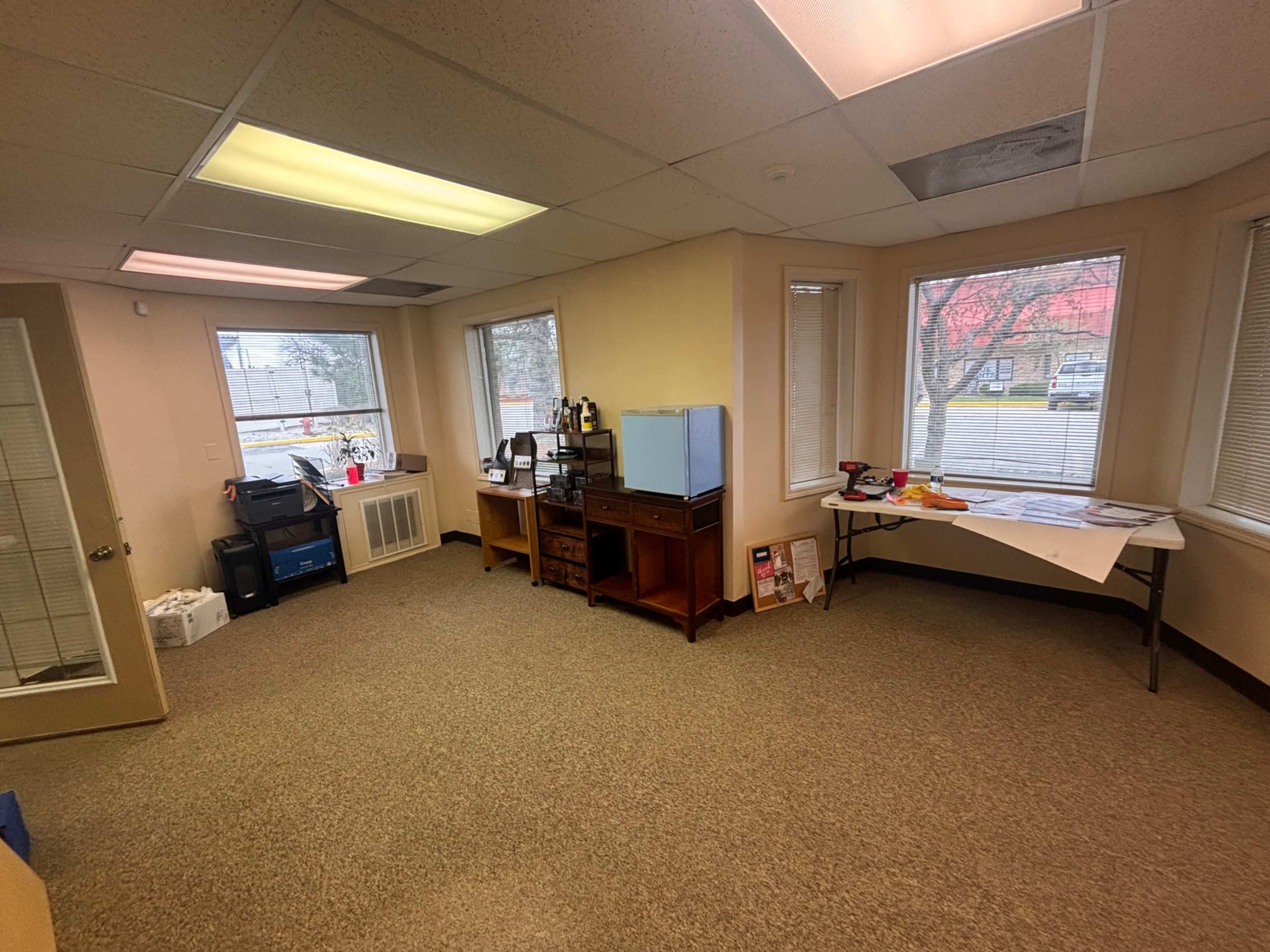 The image shows a small office space with two windows, a table on the right, a wooden cabinet, and various office supplies and equipment.