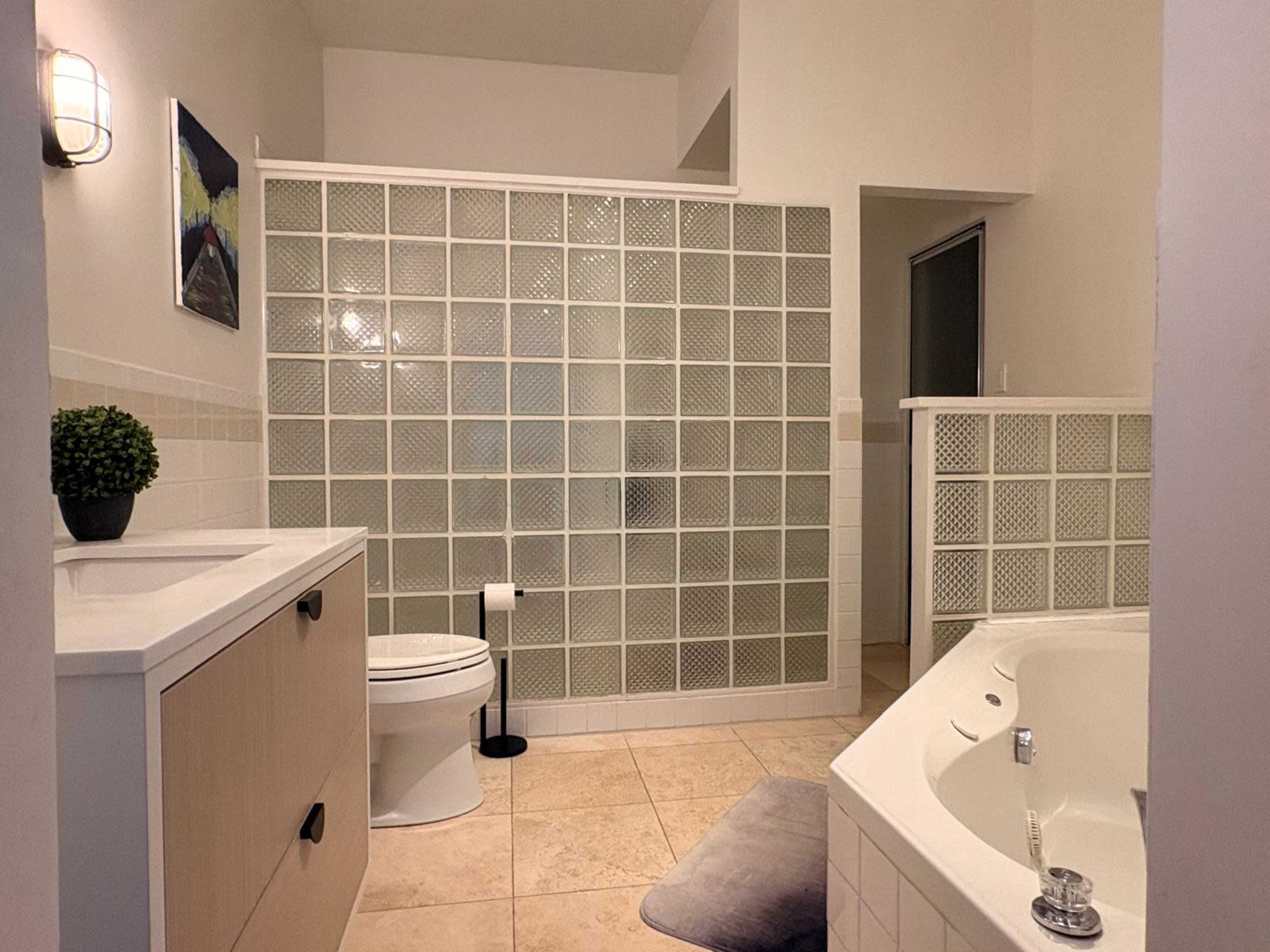 The image shows a spacious bathroom featuring a glass block partition, a freestanding bathtub, a vanity with a sink, and neutral-colored tiled flooring.