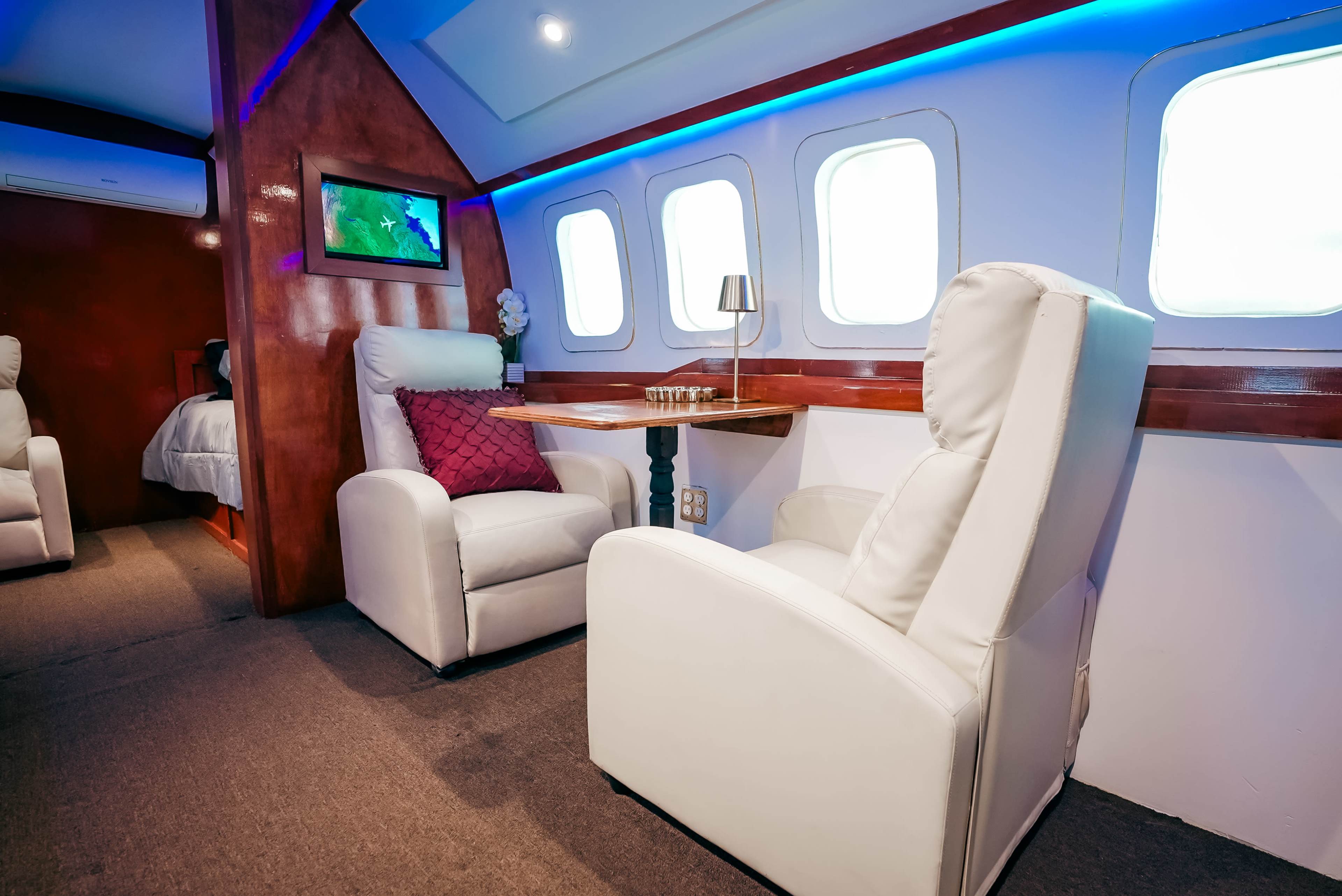 The image shows a cozy interior of a uniquely designed room resembling an aircraft cabin, featuring two white armchairs and a small table, with blue ambient lighting and large windows.