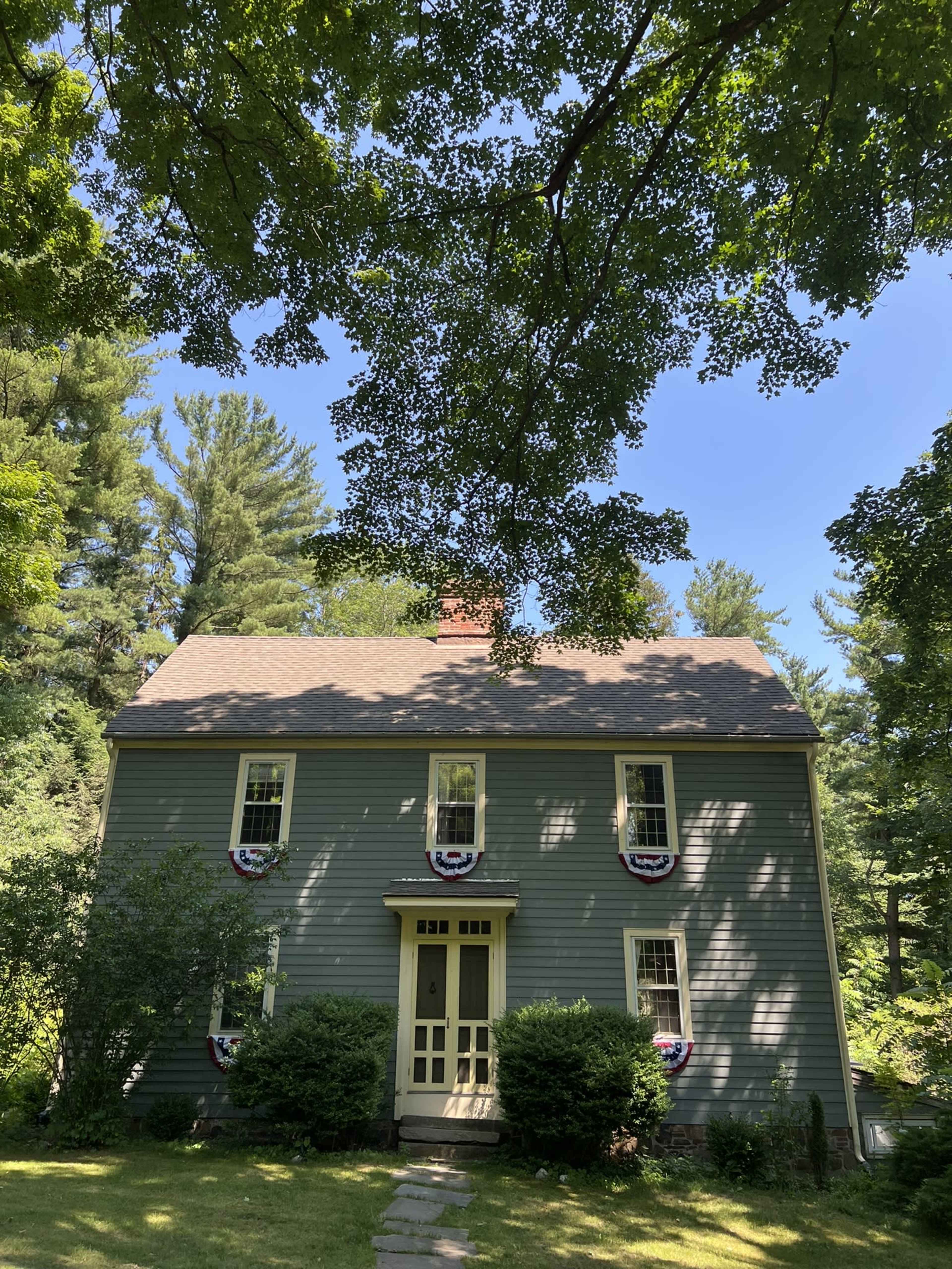 A blue wooden house with white trim and patriotic decorations is framed by trees and a clear blue sky.
