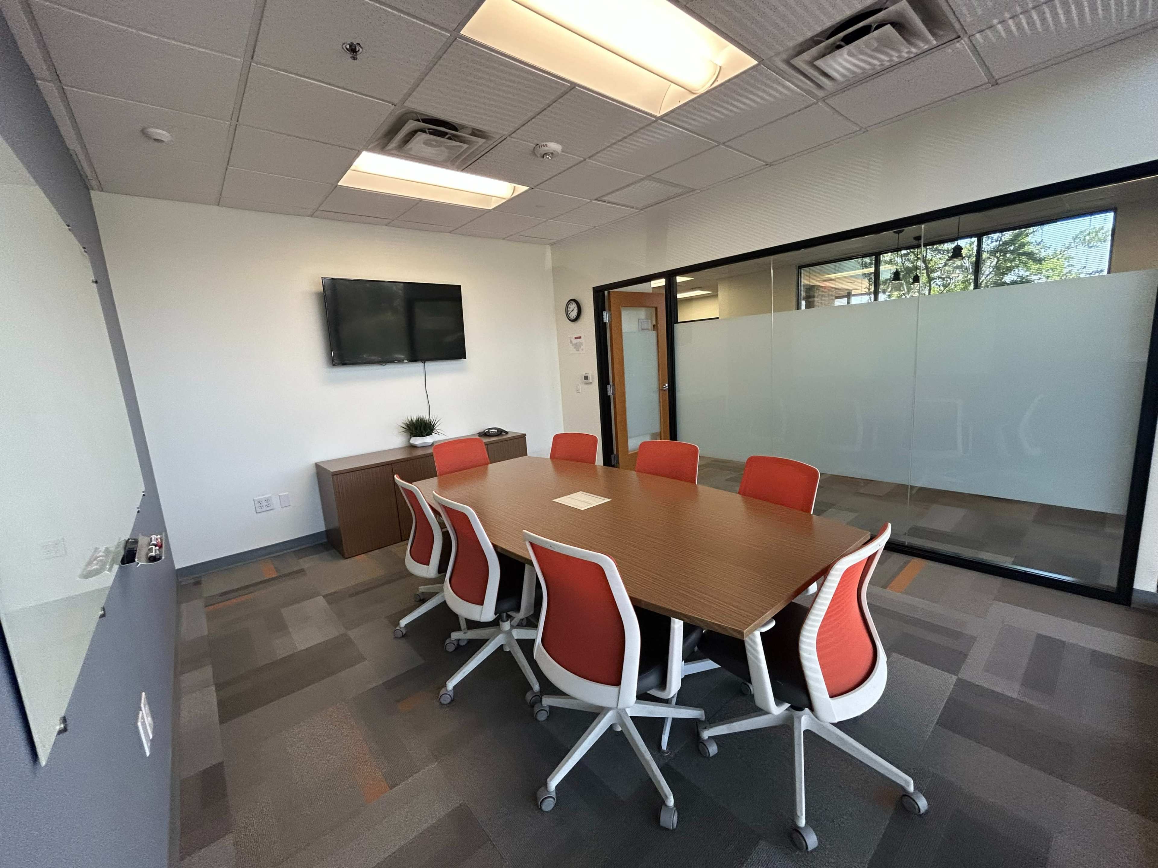 The image shows a modern conference room featuring a long wooden table surrounded by orange and white ergonomic chairs, with a television on one wall and glass walls adjacent to the space.