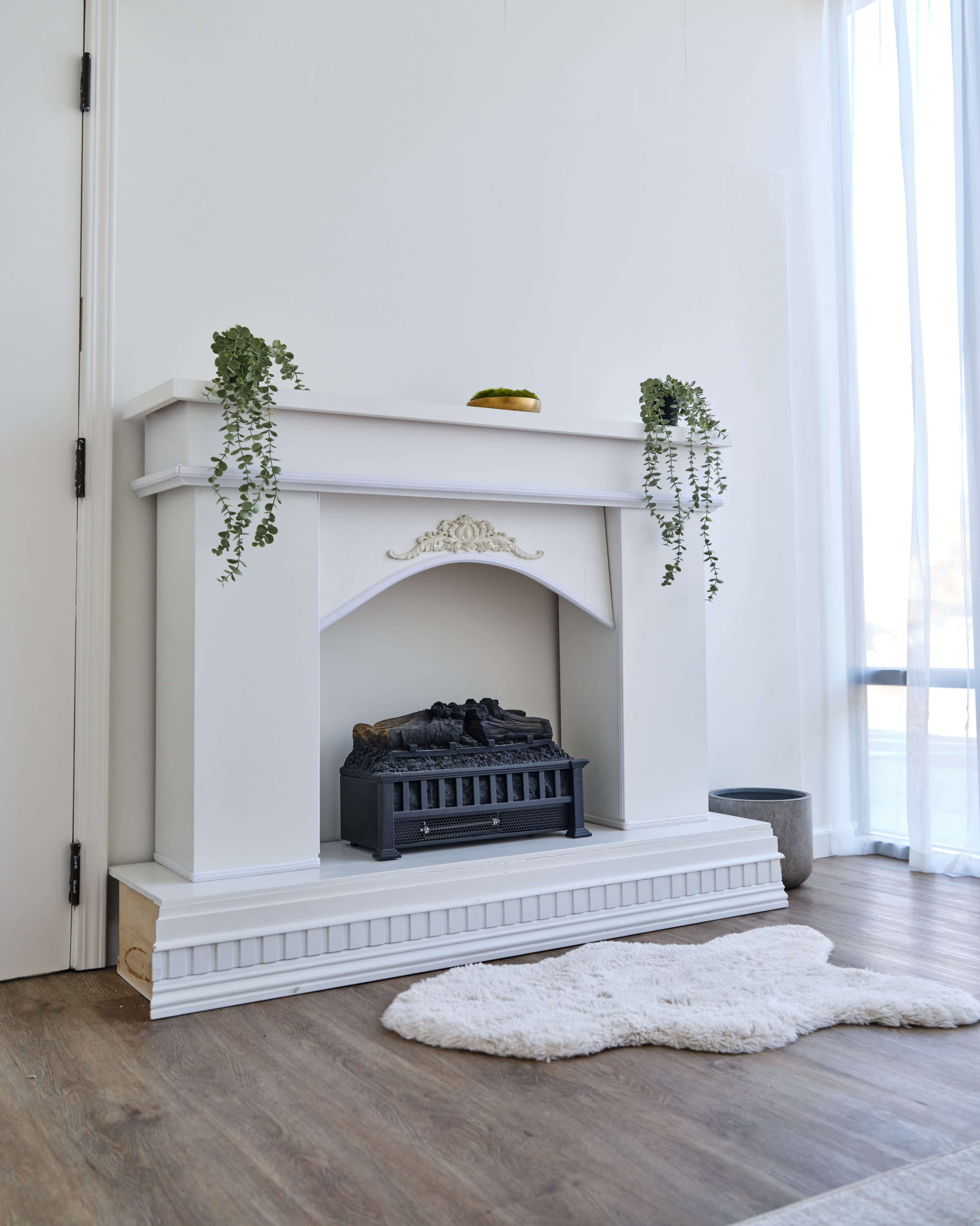The image shows a white fireplace with a decorative mantel, featuring a black electric insert and two hanging plants, set against a light-colored wall.