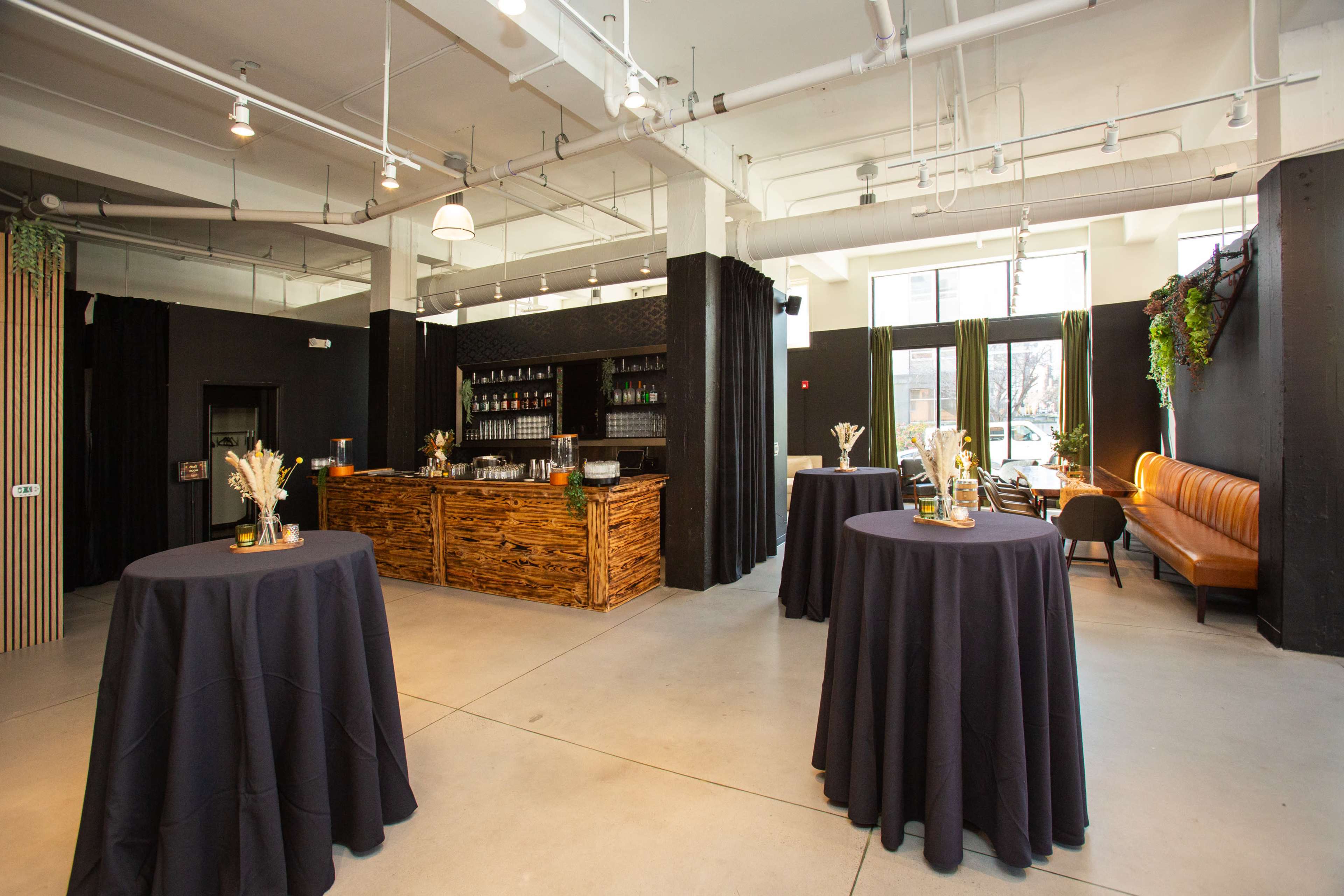 The image shows a modern event space featuring a bar with a wooden counter, several round tables with dark tablecloths, and a seating area with a leather couch.