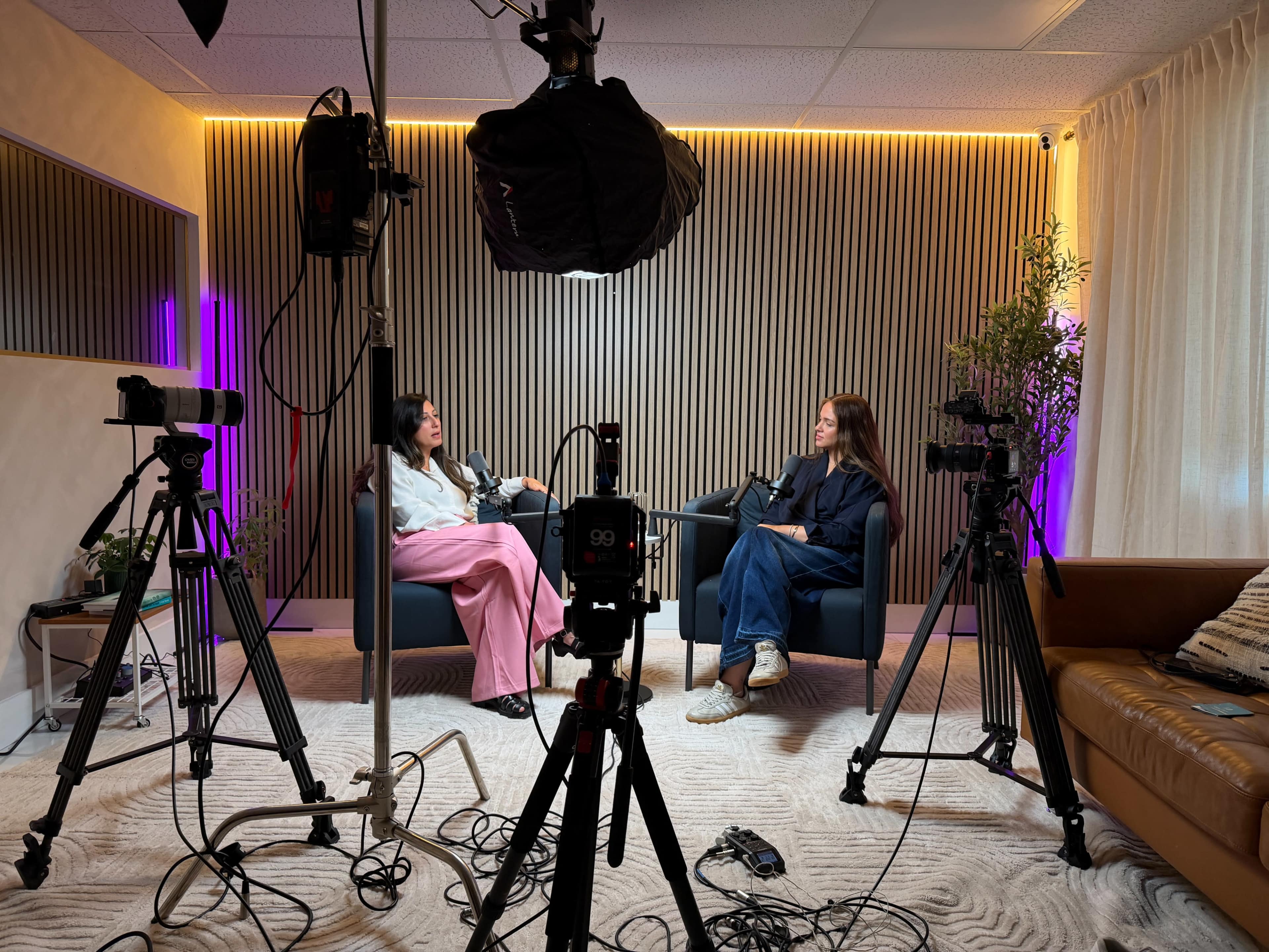 Two women are seated in armchairs in a studio setting, surrounded by cameras and lighting equipment, engaged in a conversation.