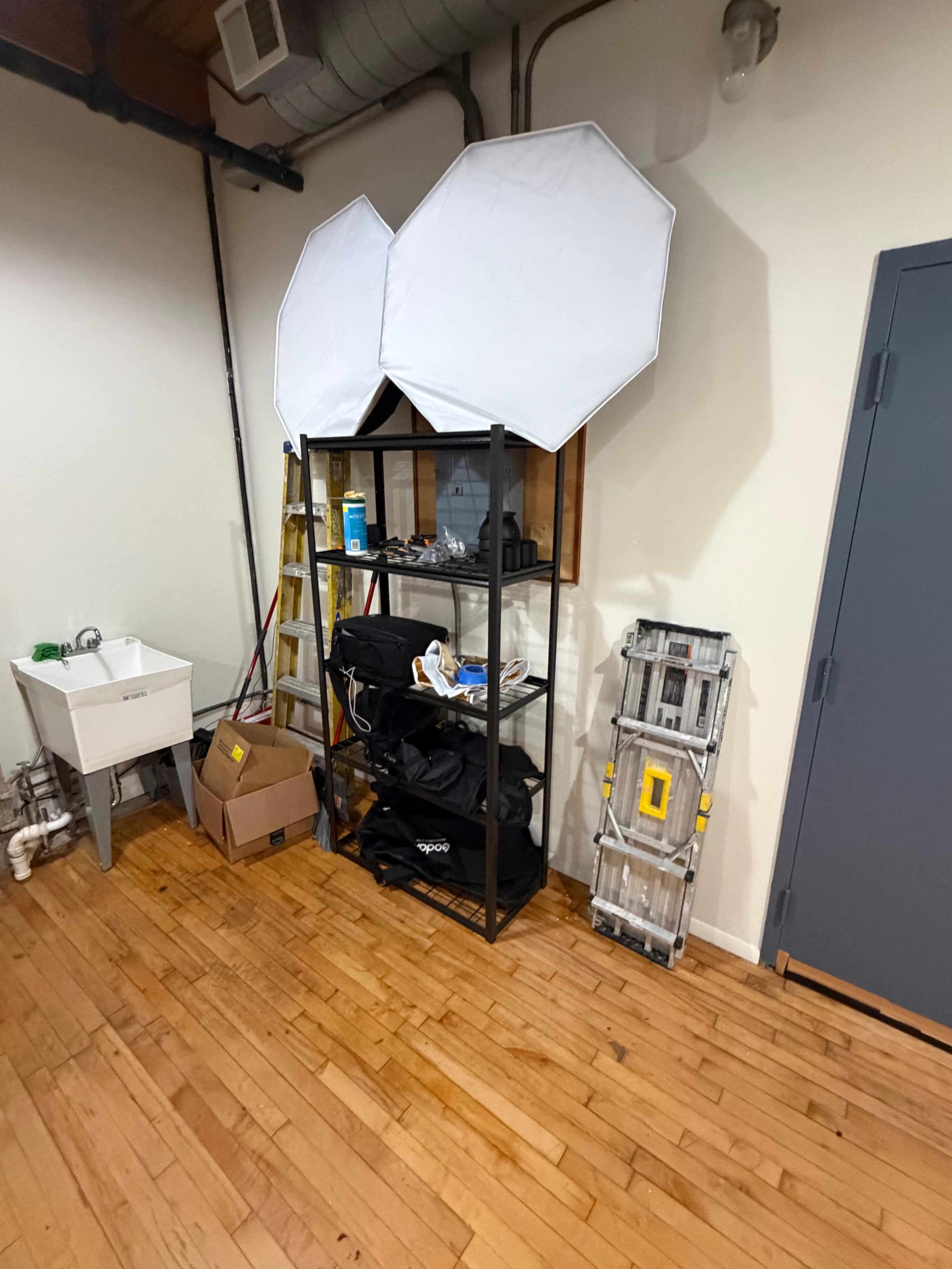 The image shows a corner of a room featuring a black shelving unit with various items, two light stands with softboxes on top, a ladder, and a white sink against the wall.