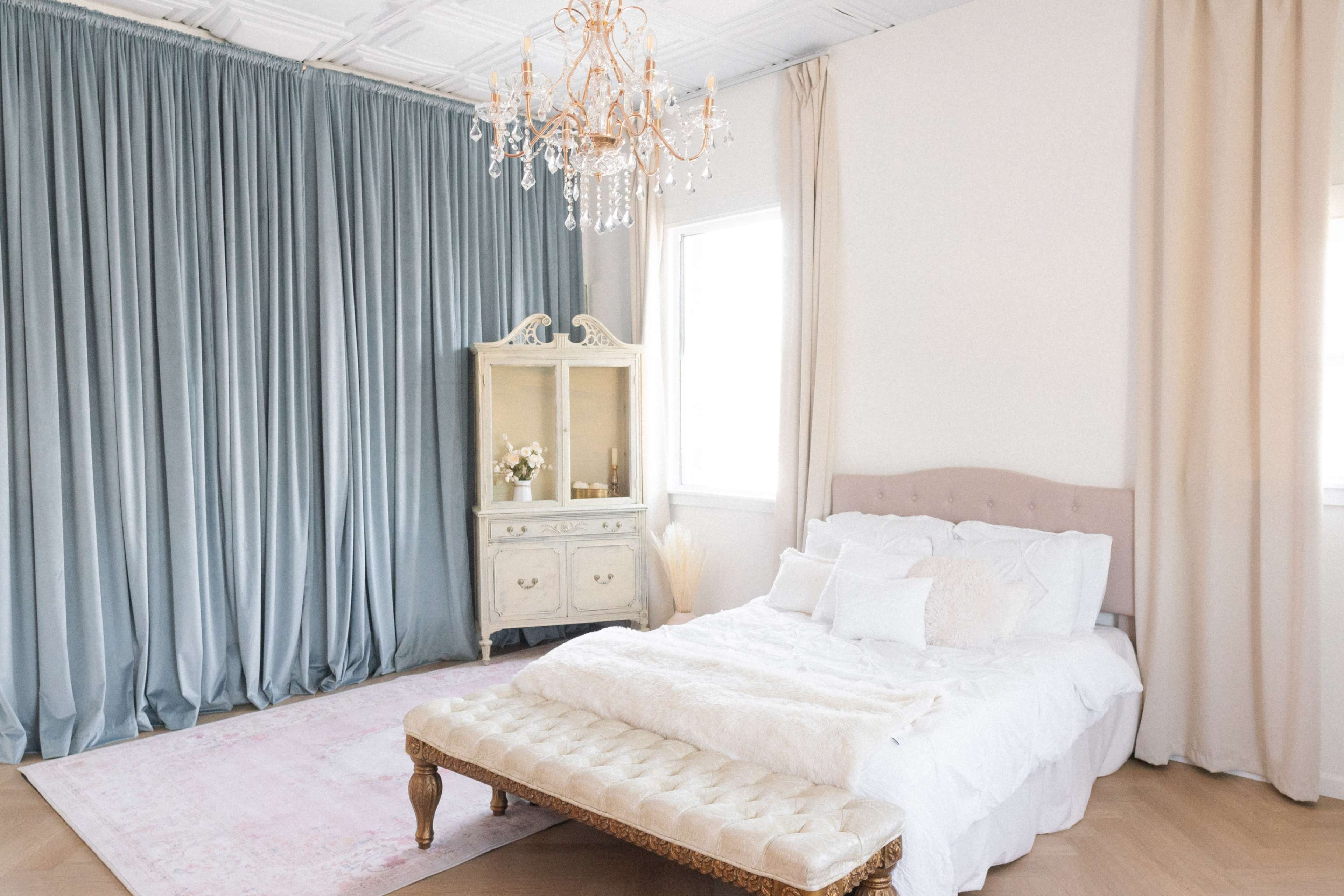 A bright bedroom features a bed with white bedding, a tufted bench, a vintage-style cabinet, and elegant curtains framing the windows.