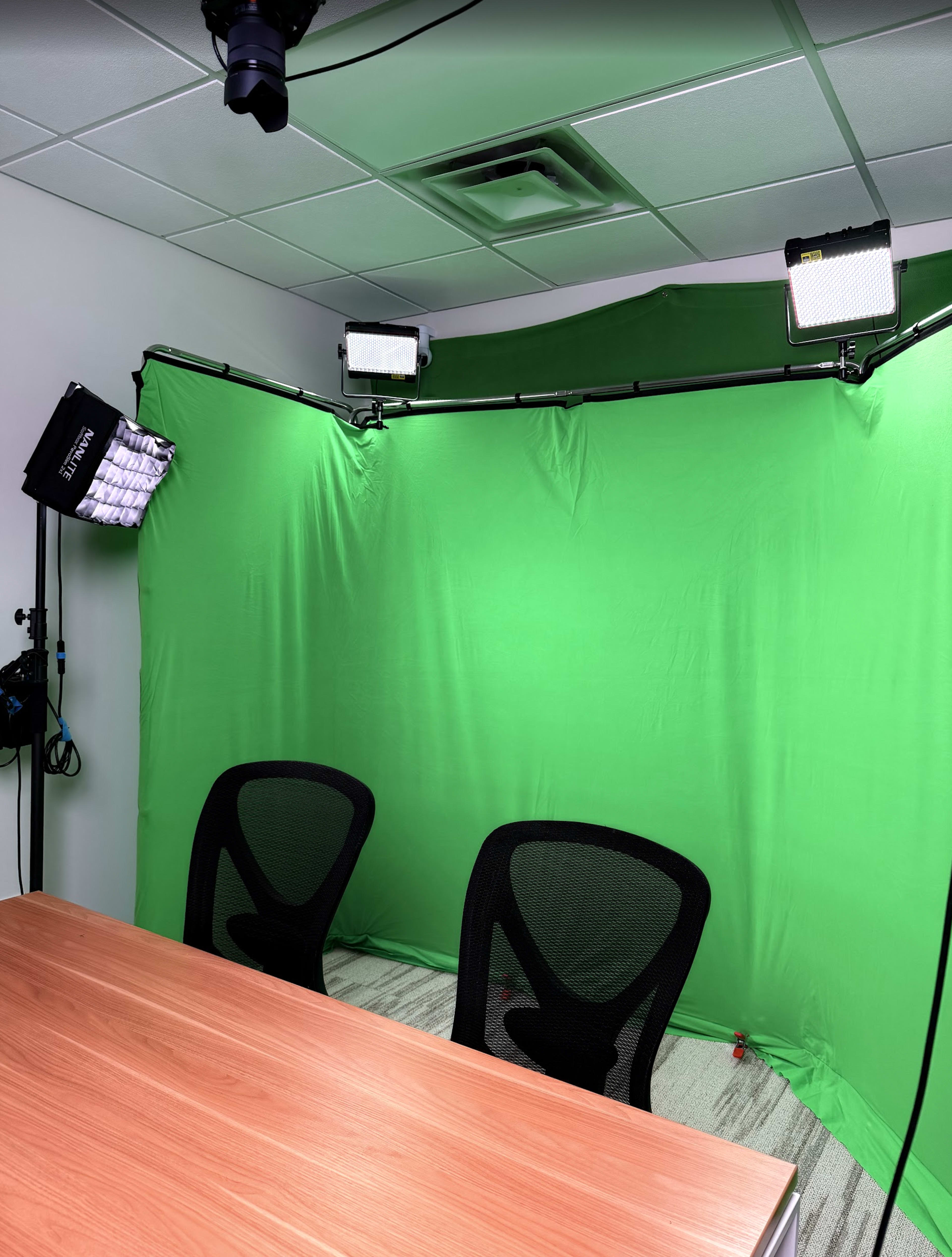 The image shows a small room setup with two ergonomic chairs facing a wooden table, and a green screen backdrop illuminated by two softbox lights.