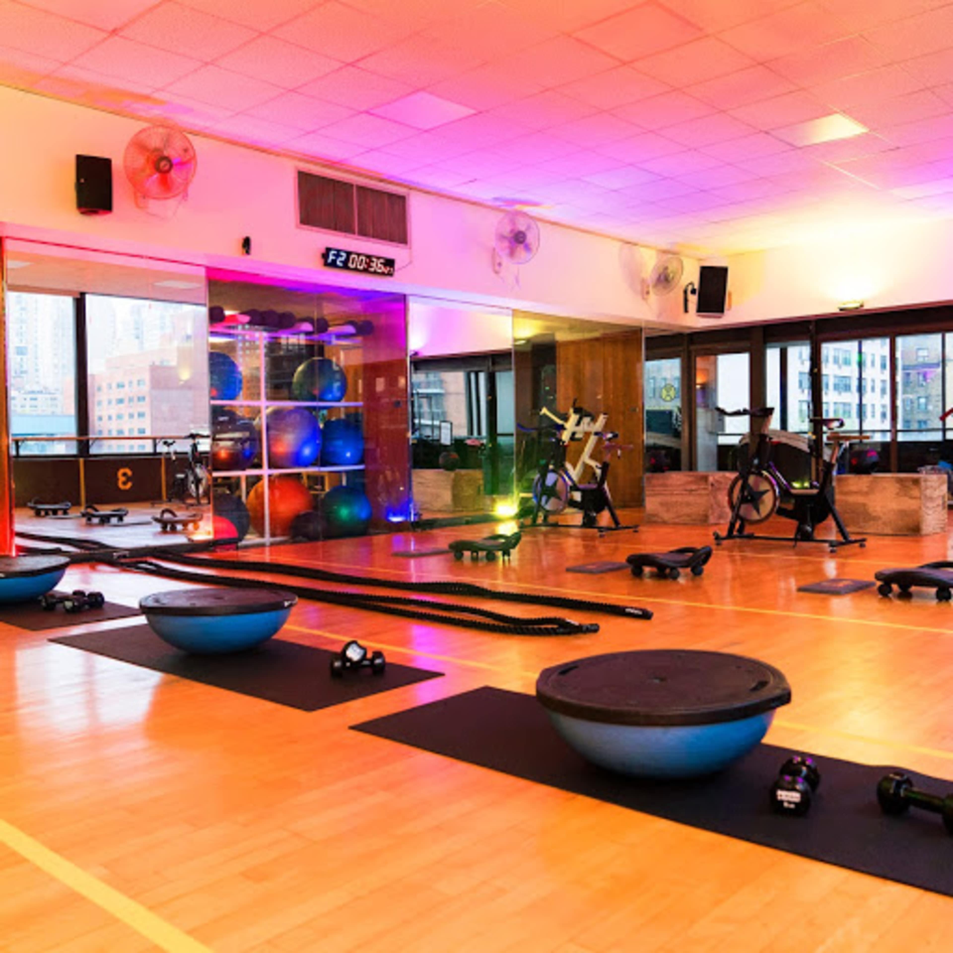 A gym interior equipped with various exercise equipment, including weights, exercise balls, and fitness machines, under colorful lighting.