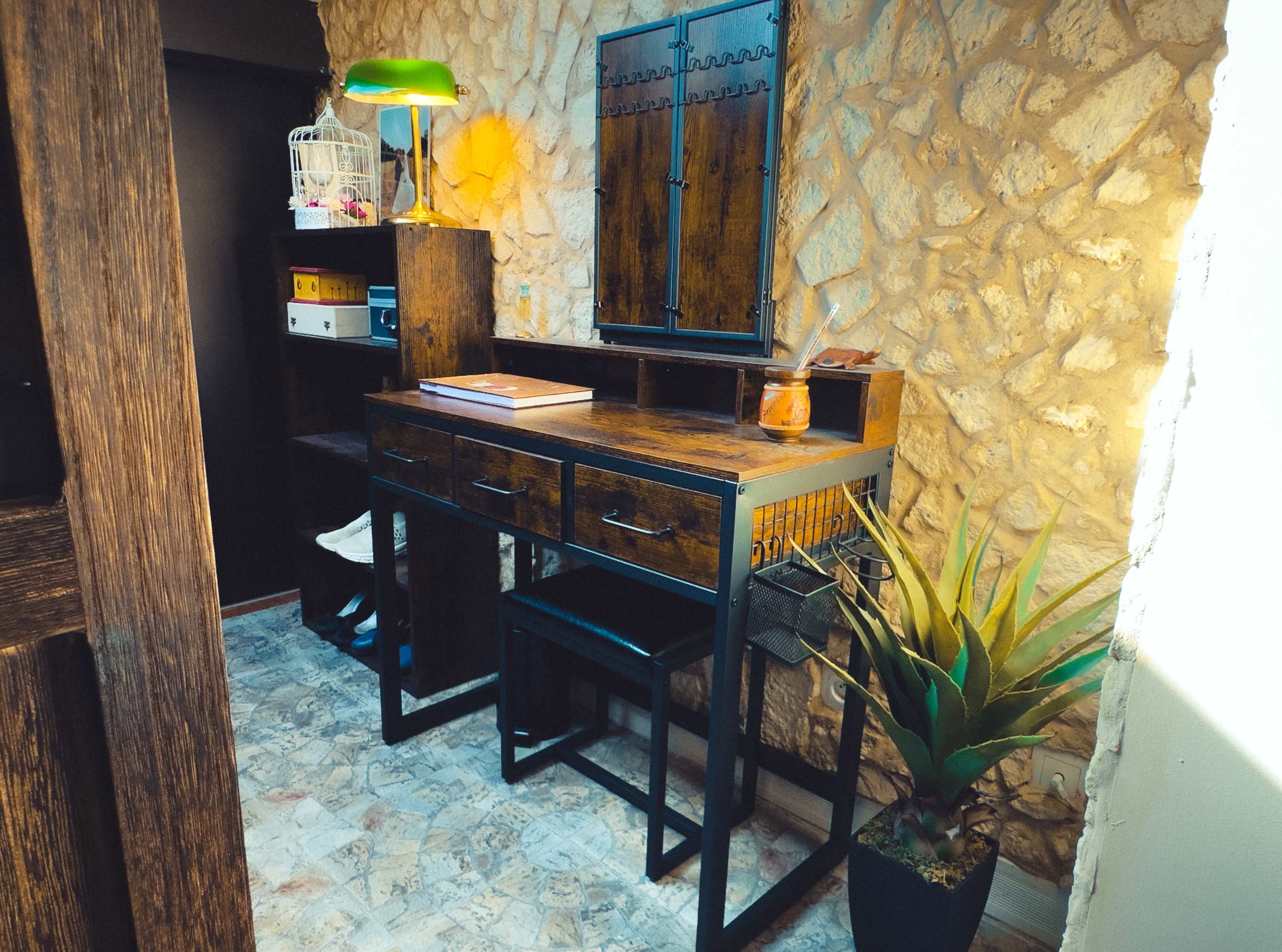 A wooden desk with three drawers and a green lamp sits against a stone wall, accompanied by a potted plant and shelves with storage boxes.