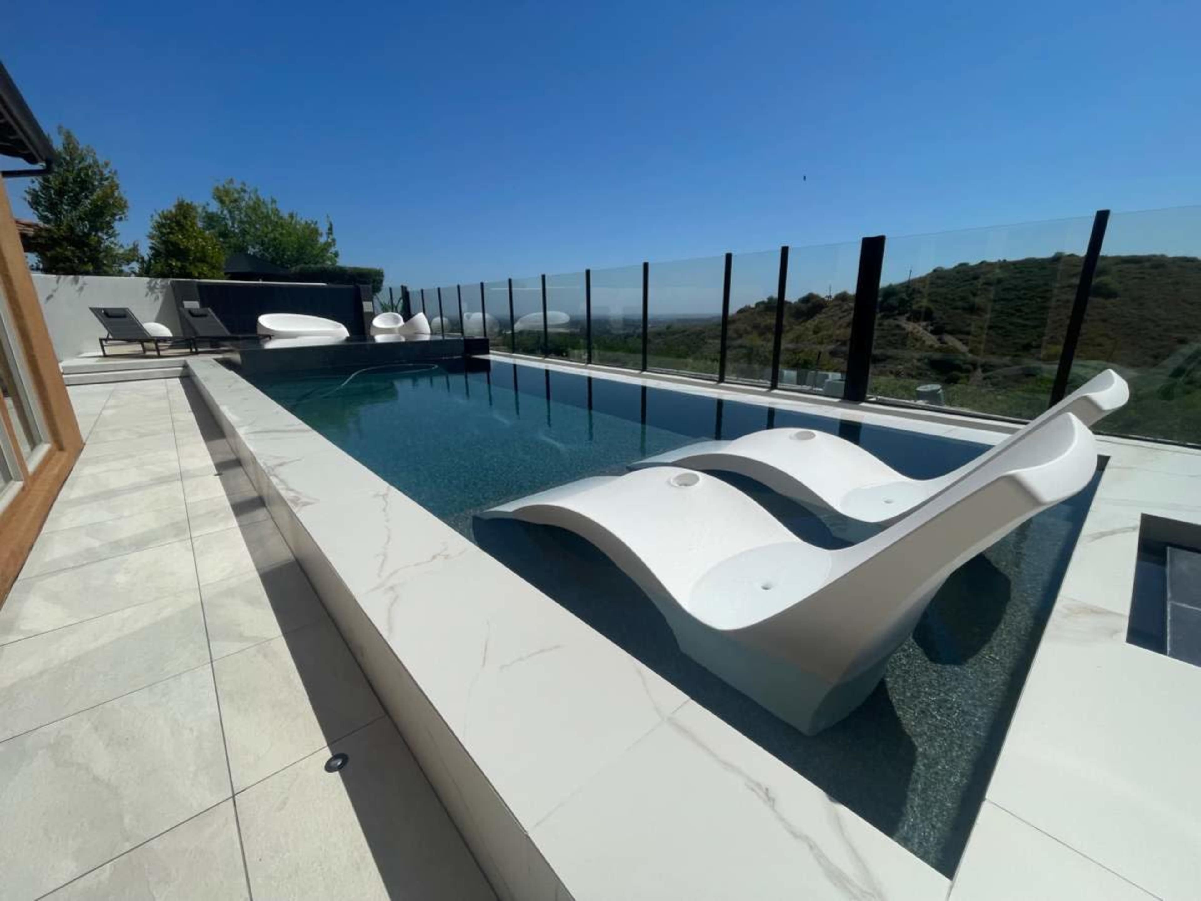 A modern swimming pool with white lounge chairs and a glass railing overlooking a scenic landscape.