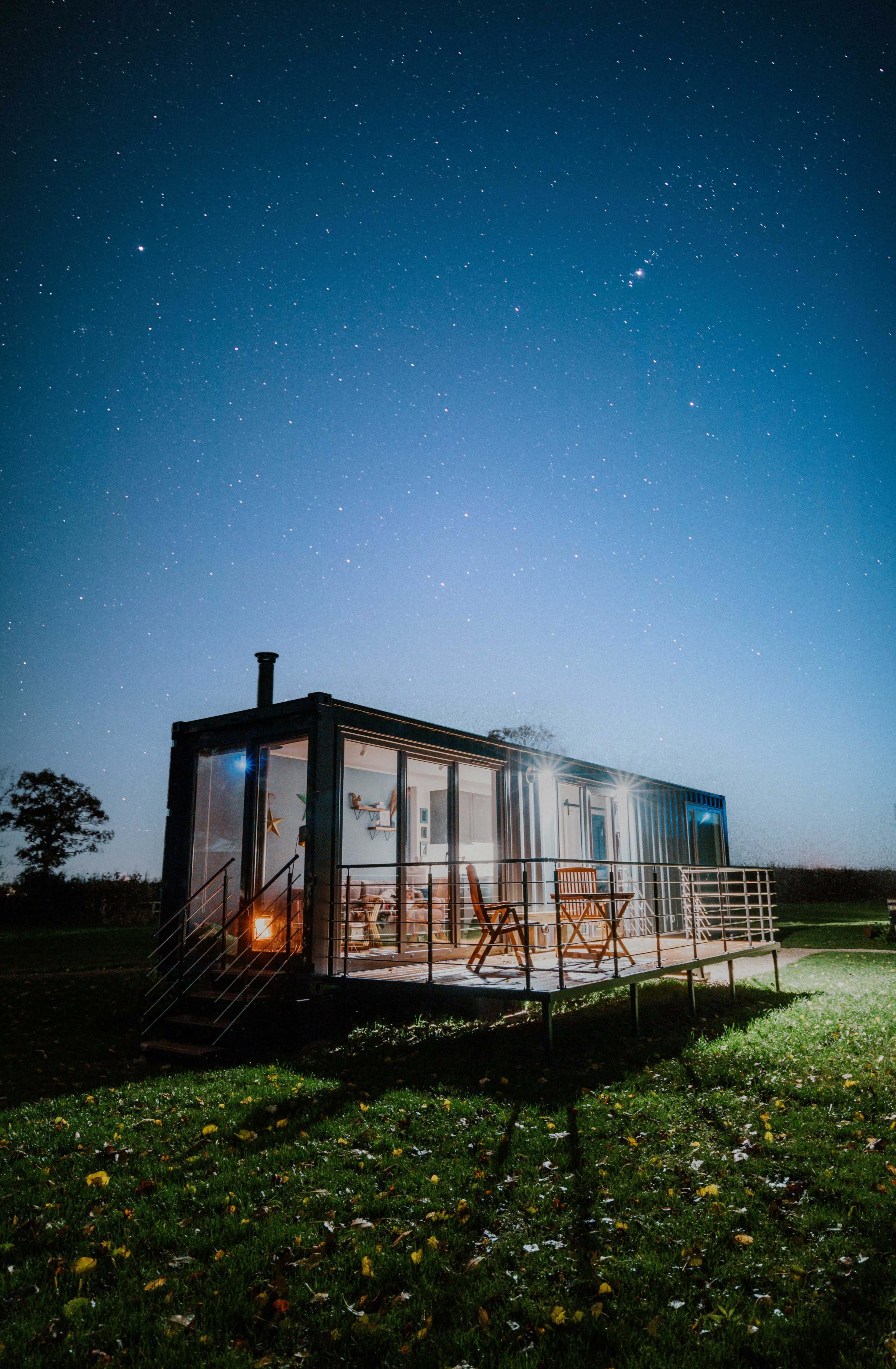 A modern glass cabin sits in a grassy field under a starry night sky.