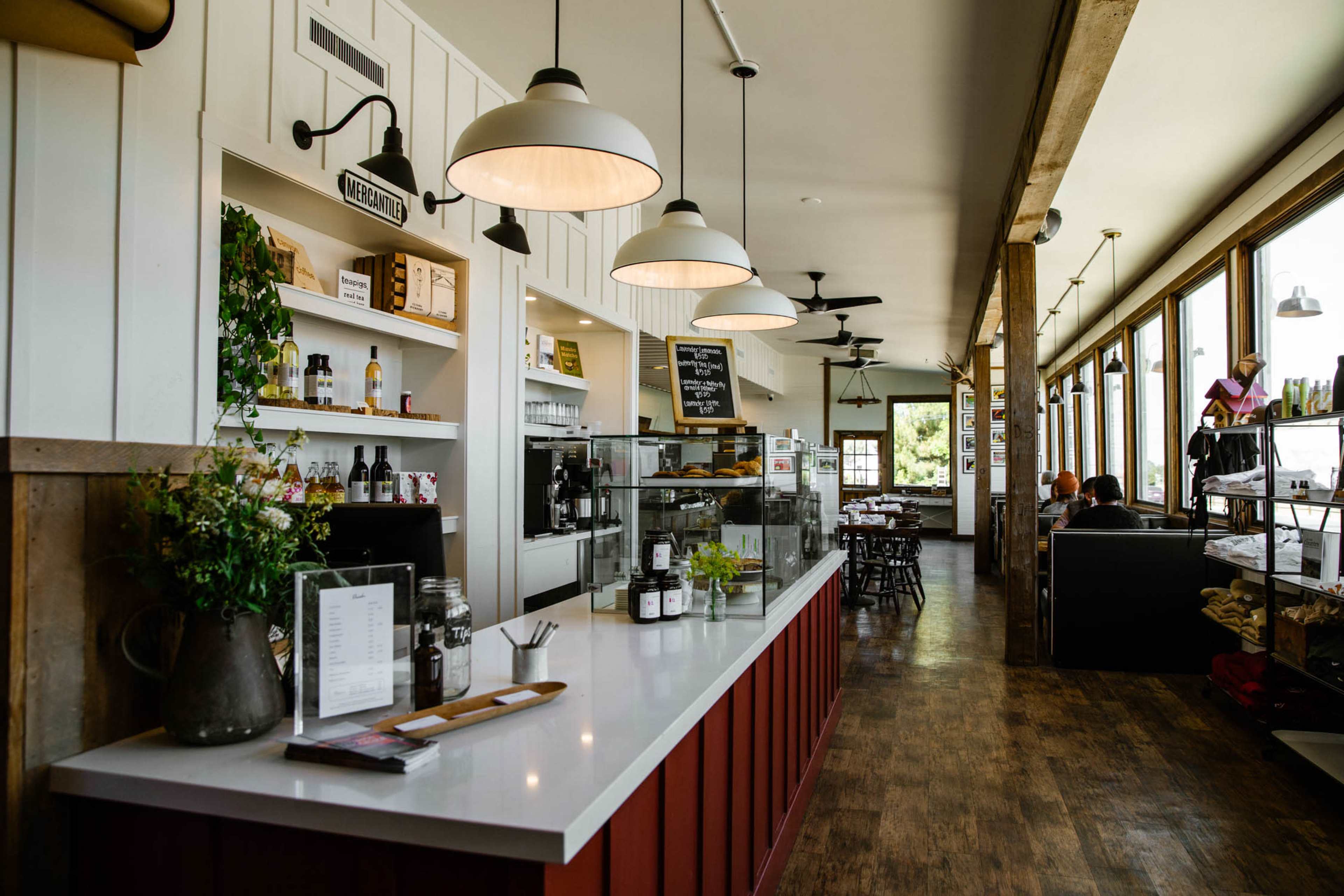 A cafe interior featuring a service counter with a display case, shelves of products, and tables along large windows.