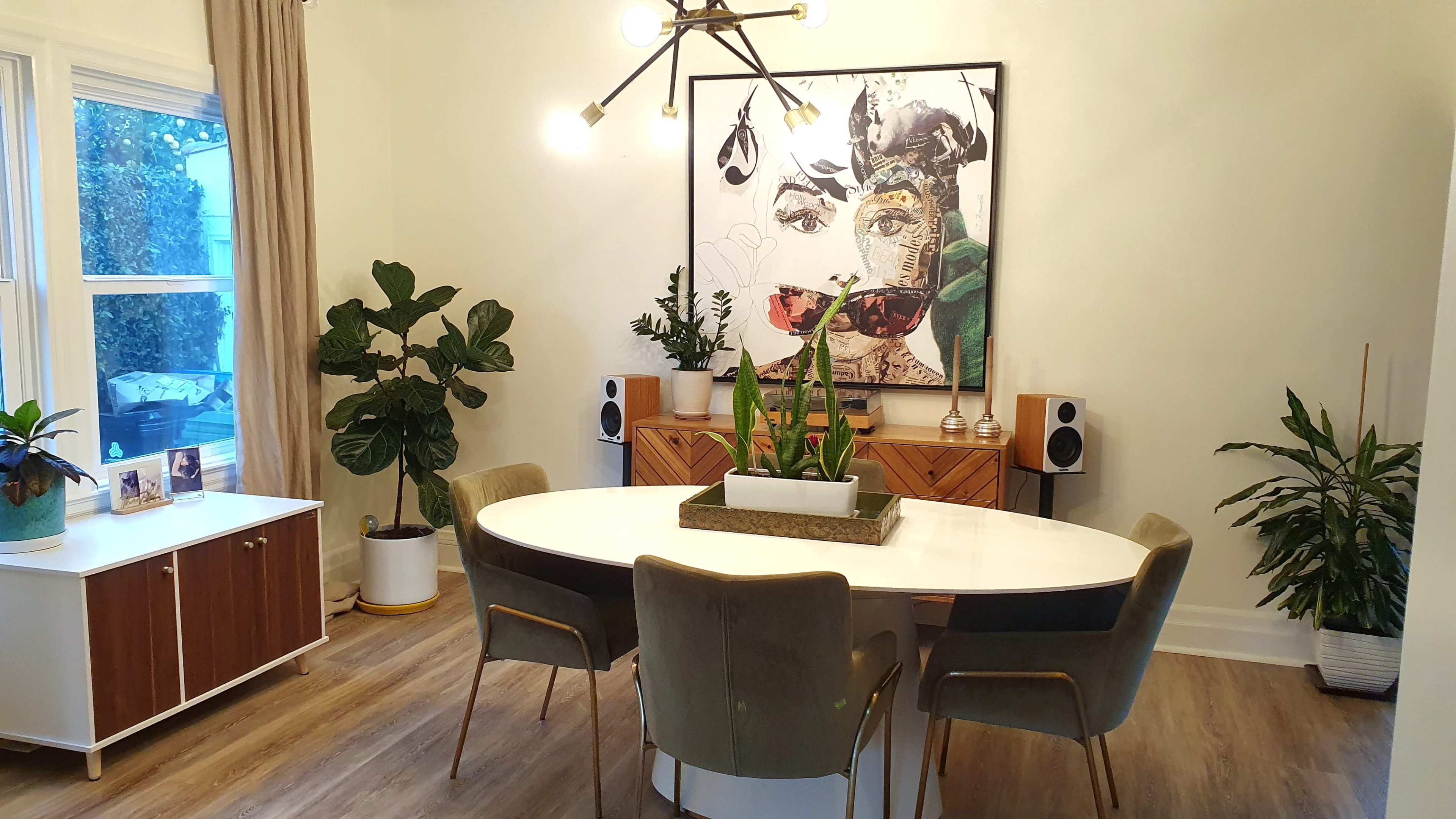A dining area features a round white table surrounded by green chairs, with a large artwork on the wall and various plants positioned around the space.